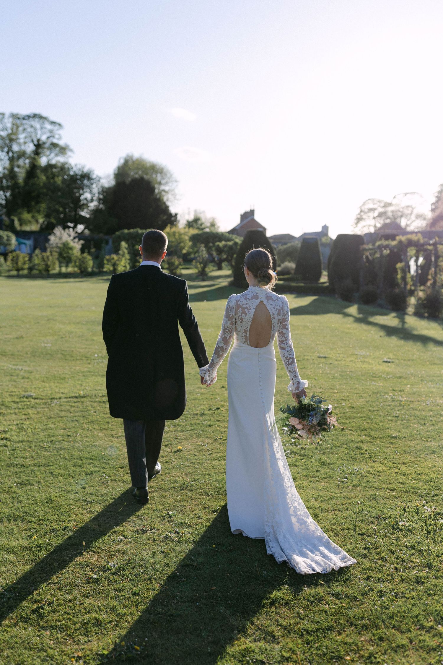 A couple walks hand in hand across a sunlit grassy field casting long shadows during golden hour.