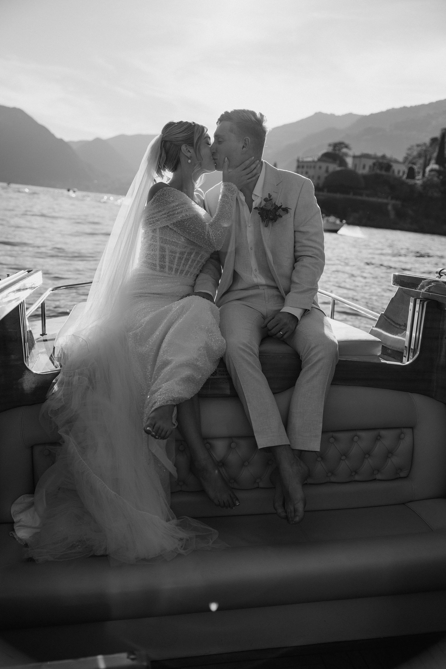 Black and white photo of a bride and groom kissing on a boat with mountains and lake in the background.