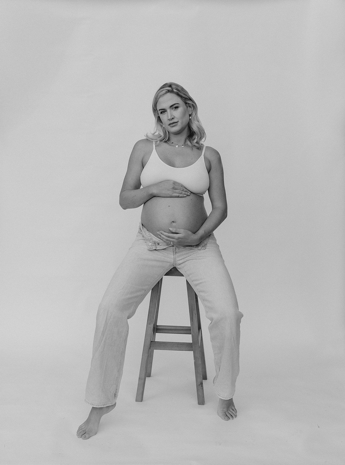Black and white maternity photo of a pregnant woman in a crop top and jeans sitting on a stool.