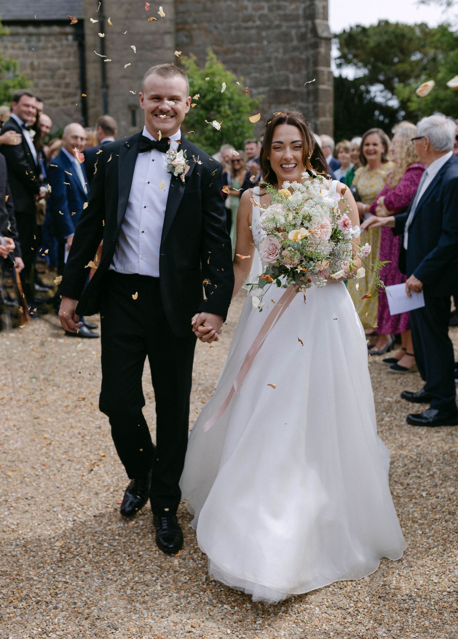 Wedding party walks down gravel path while guests throw confetti in celebration.