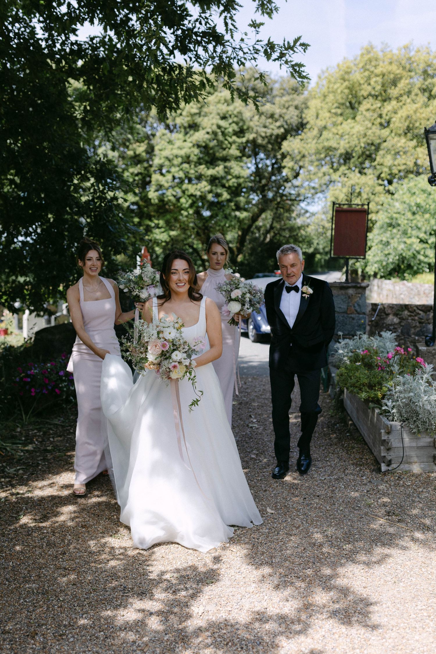 Wedding party walking along a tree-lined gravel path in elegant white dresses.