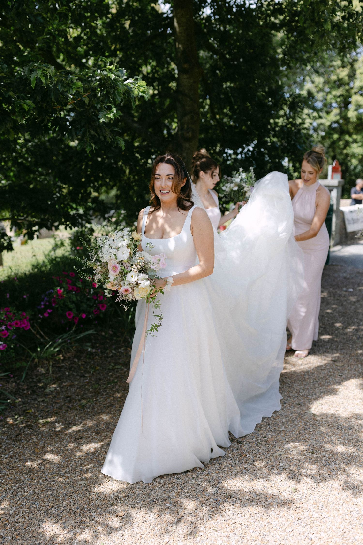 Wedding party members in white dresses walk along a sunlit garden path carrying bouquets.