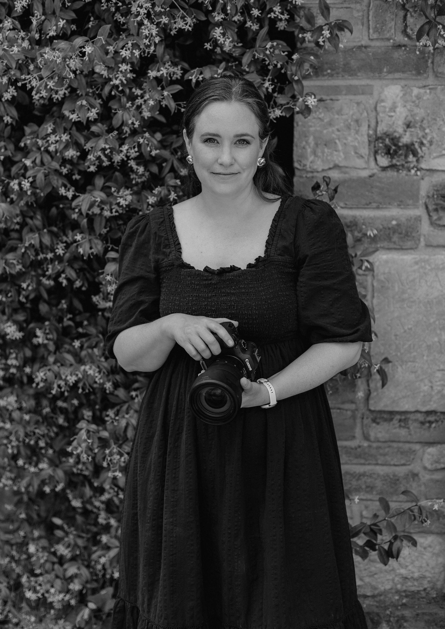Professional photographer in a black dress poses with camera against an ivy-covered stone wall.