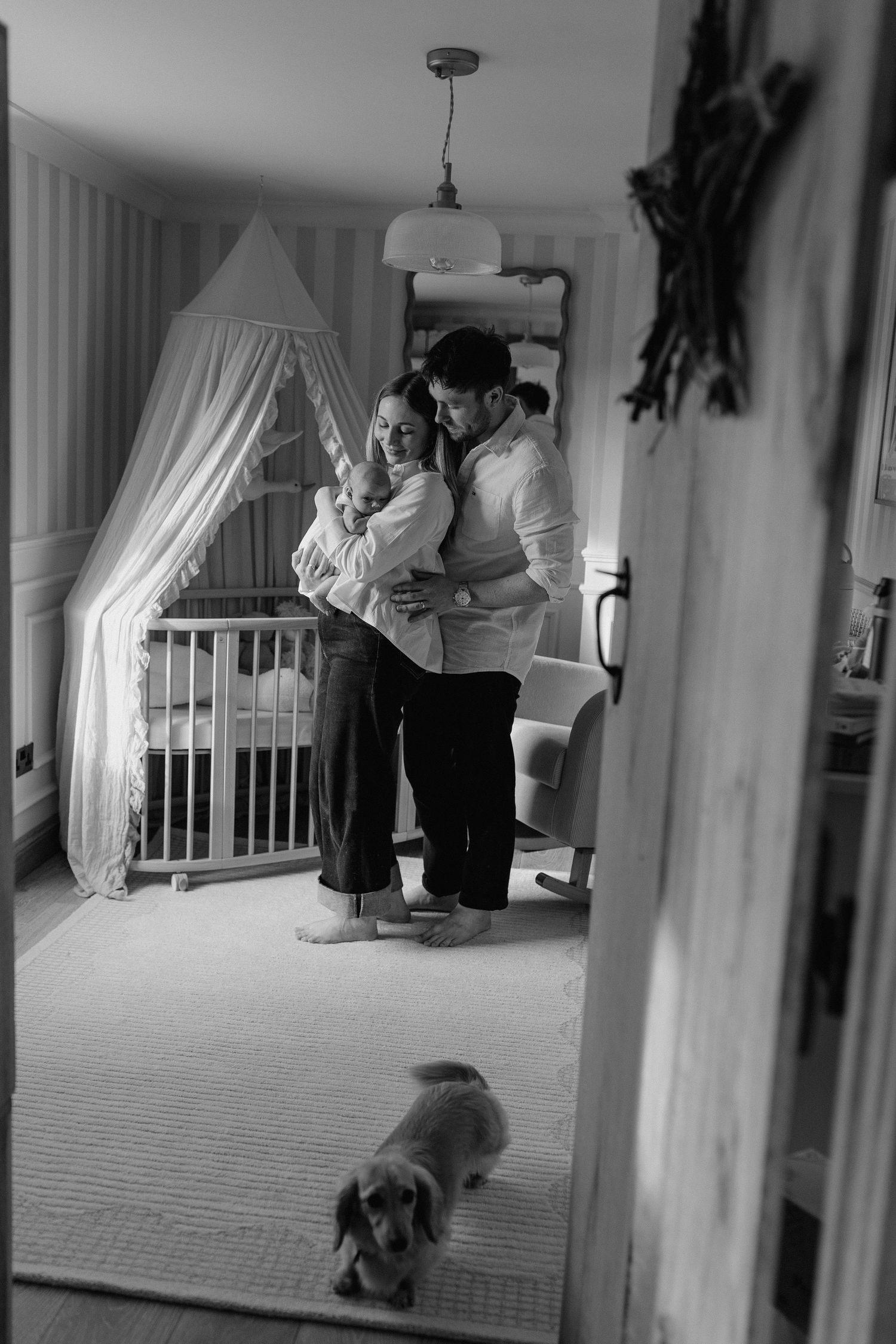 Black and white photo of couple holding newborn baby in nursery with canopy crib and dog in foreground.
