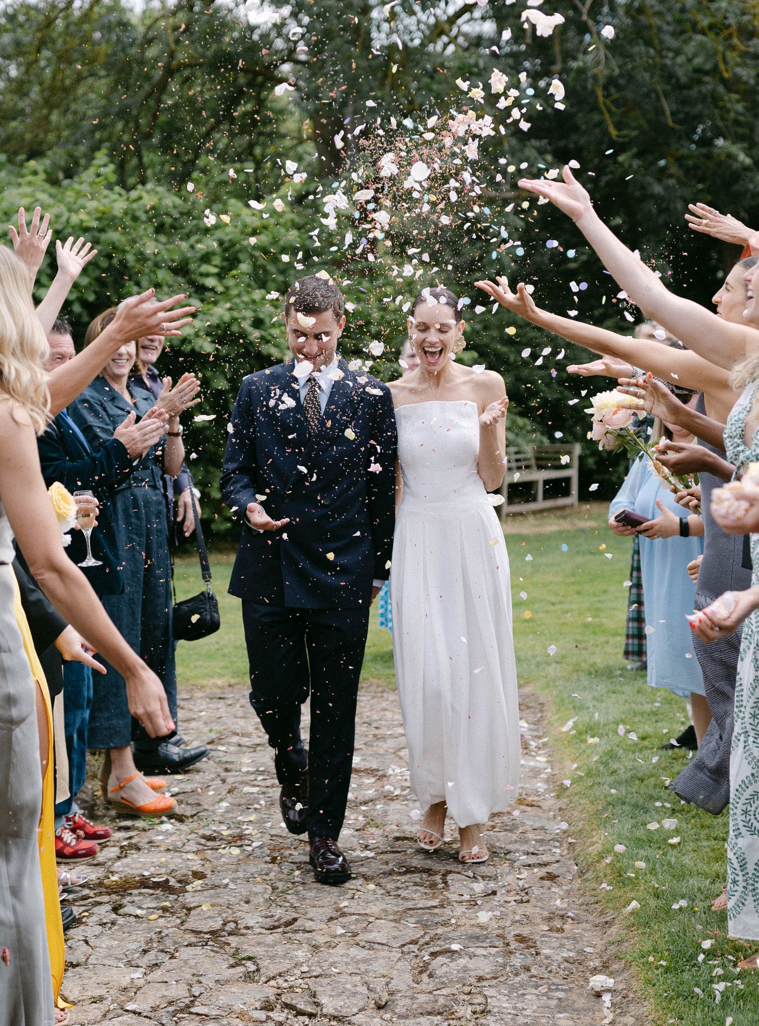 Wedding guests throw confetti in celebration as they line a pathway outdoors.