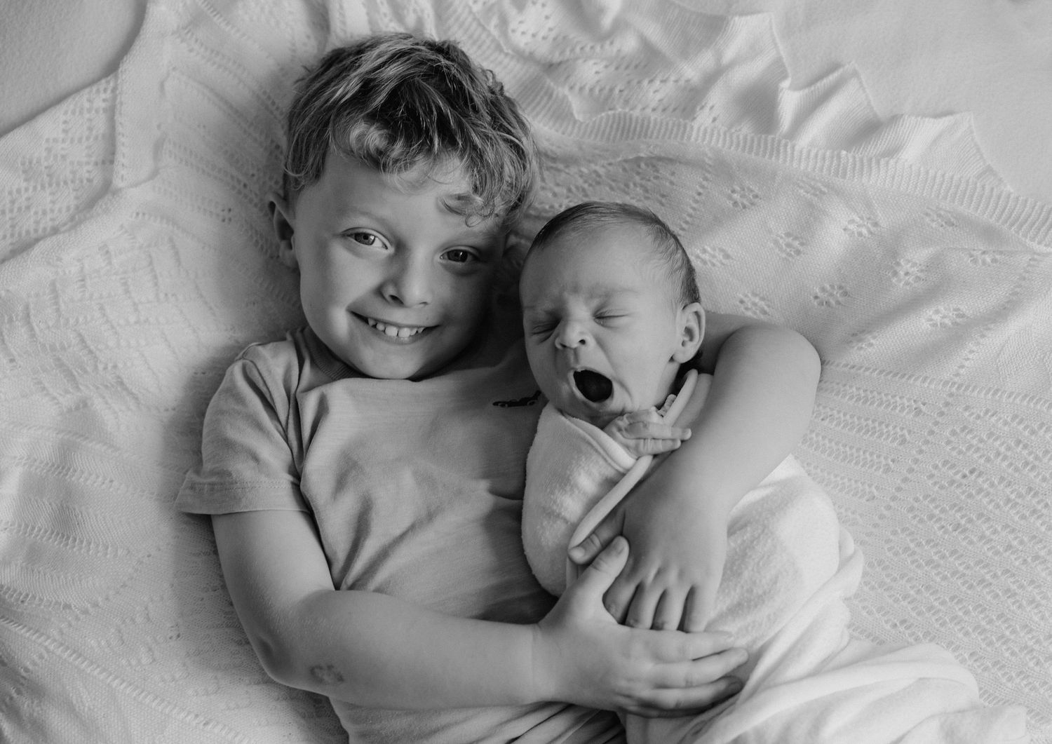 Black and white photo of a smiling boy lying down holding a yawning newborn baby sibling on a white blanket.