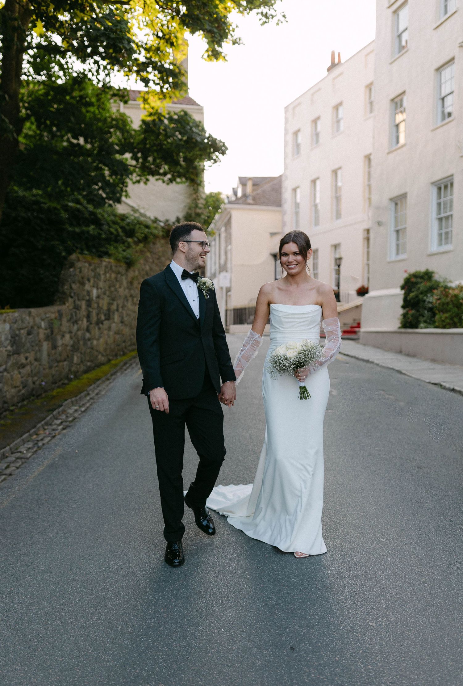 A couple walks together down a quiet street lined with white buildings on their wedding day.