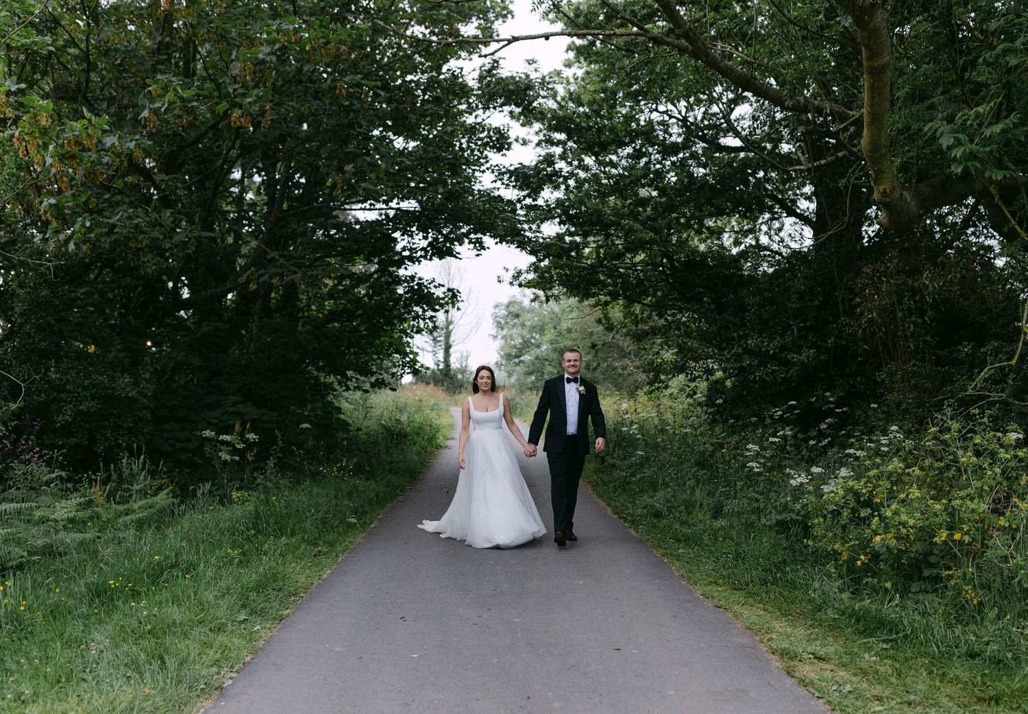Wedding couple walking down a tree-lined path surrounded by lush greenery.