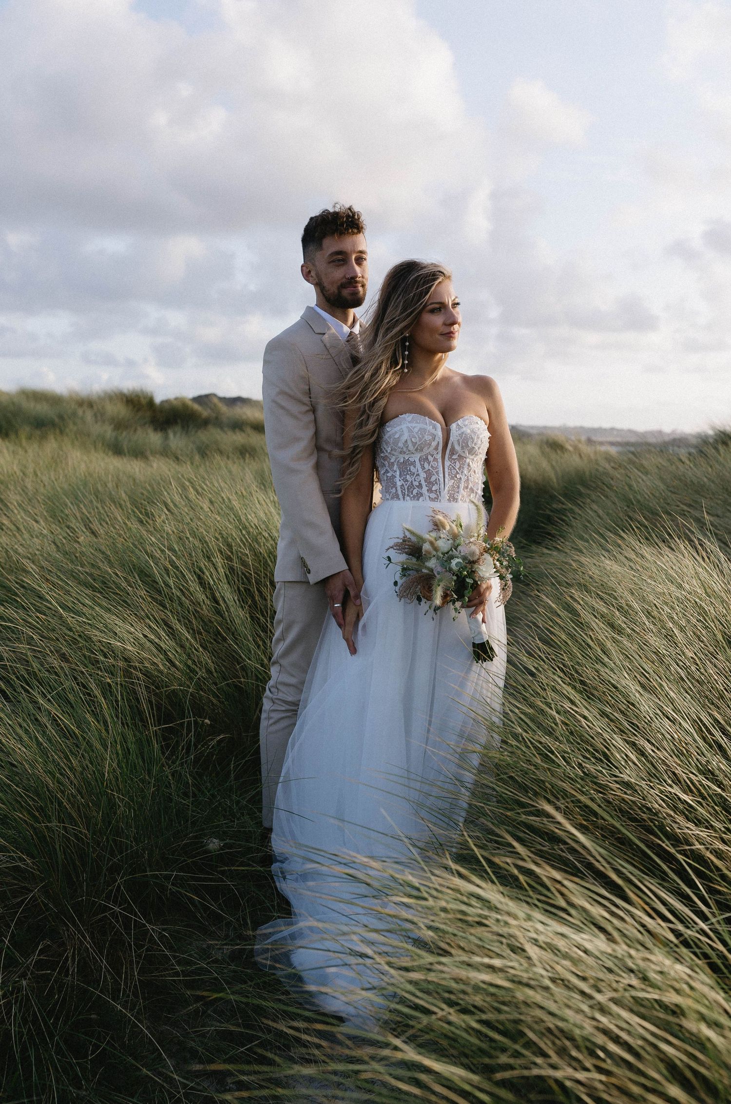Newlyweds pose among windswept beach grass during their coastal wedding photoshoot.