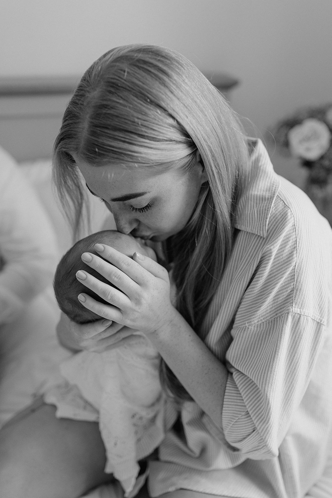 Black and white photo of a young mother tenderly kissing her newborn baby's head while cradling them gently.