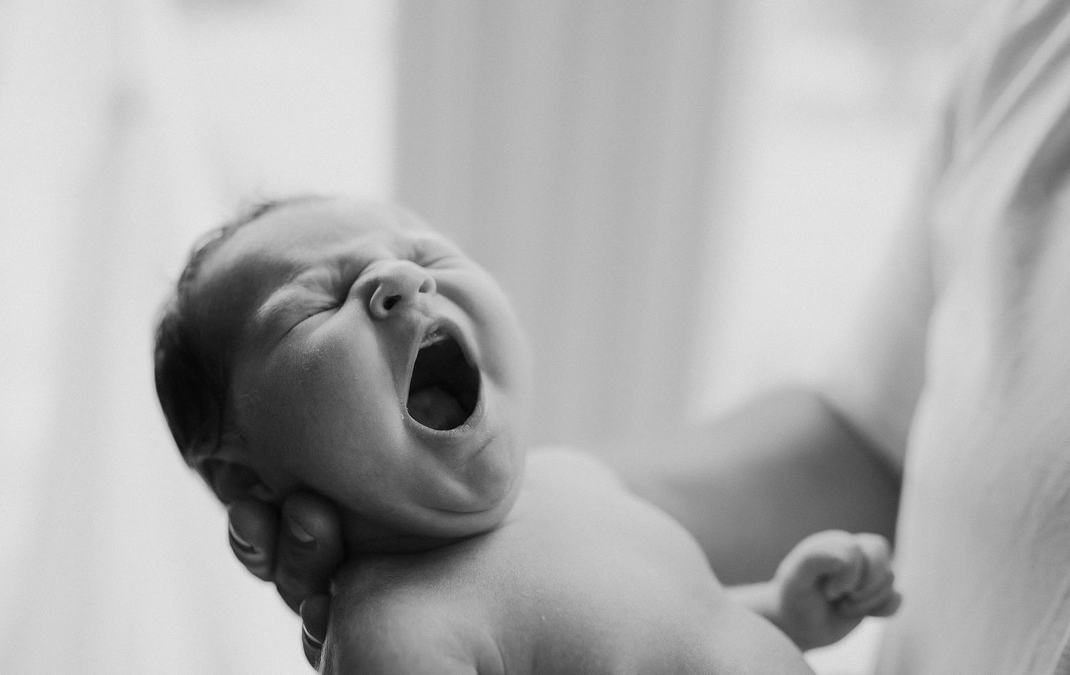 Black and white photo of a newborn baby yawning while being held, capturing a precious early life moment.