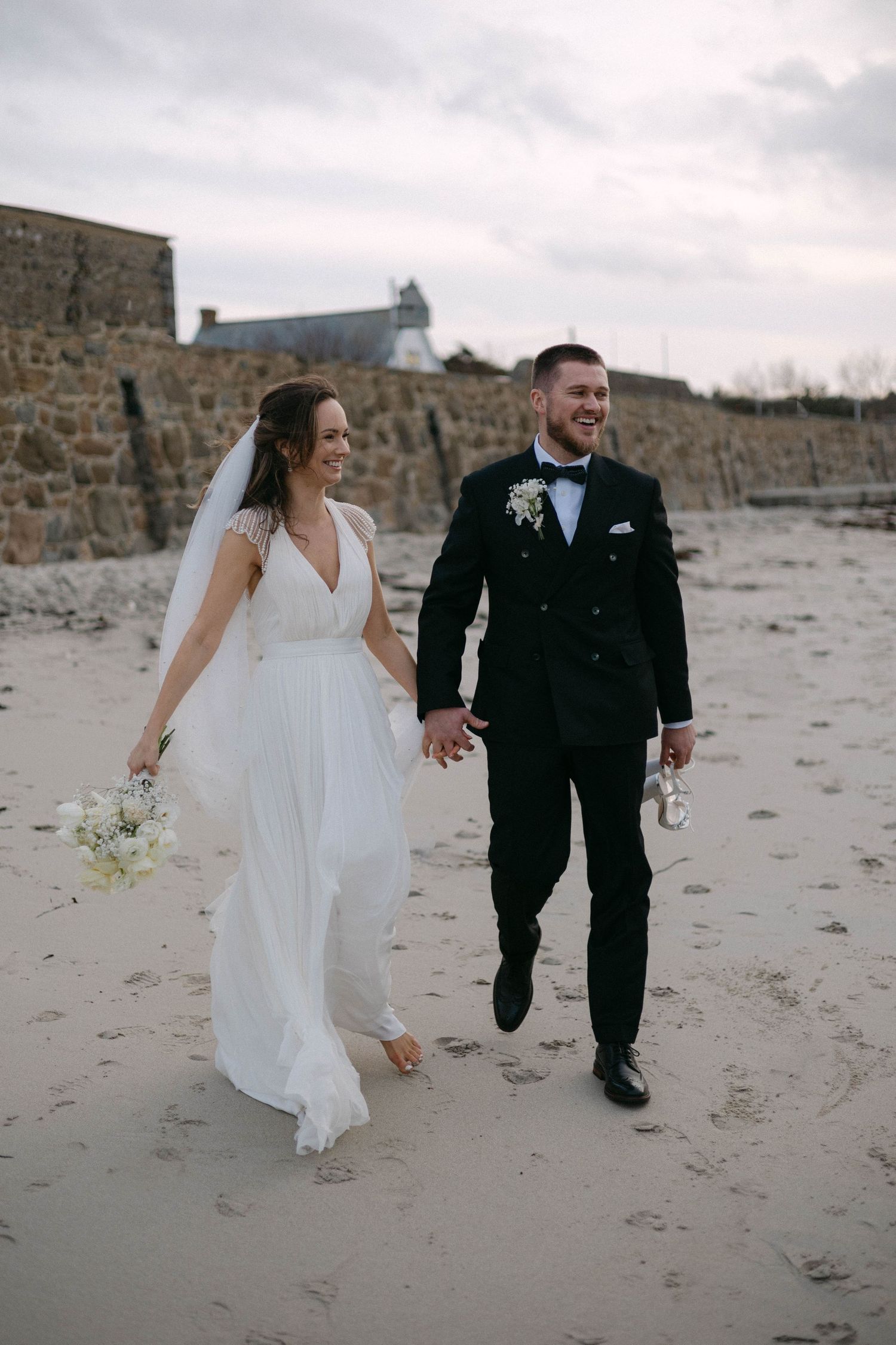 A couple strolls along a sandy beach with a stone wall backdrop.