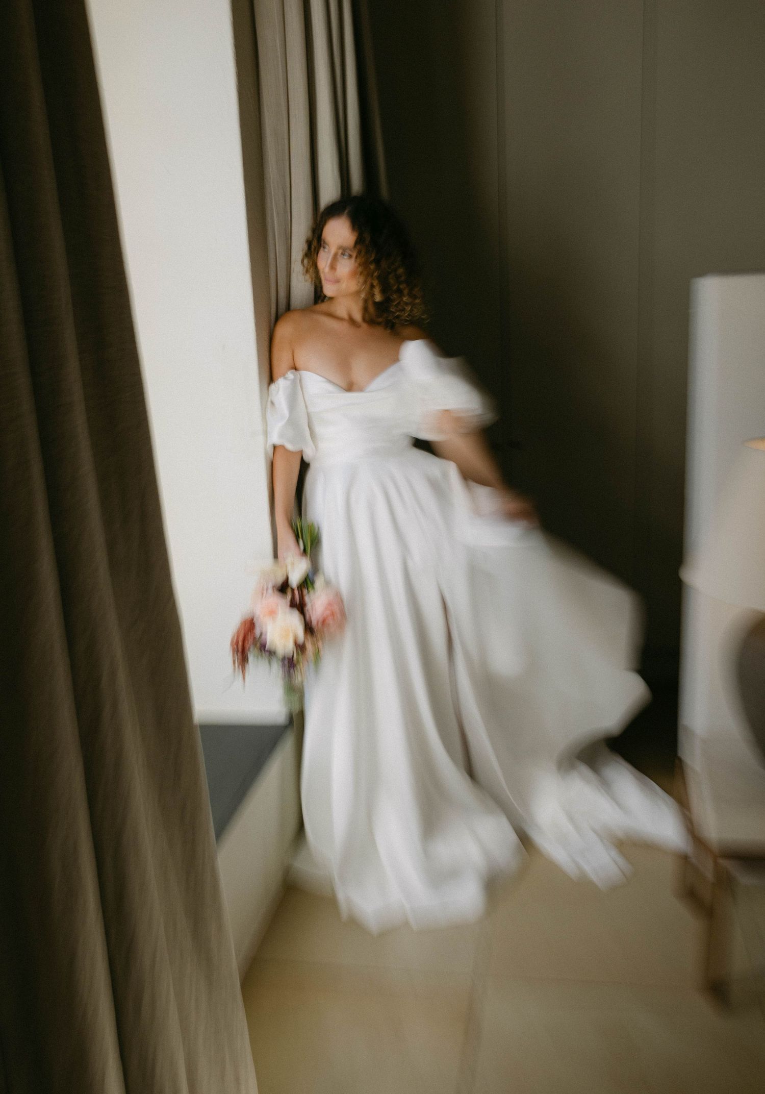 Flowing white wedding gown captured in motion against dark curtains and natural window light.