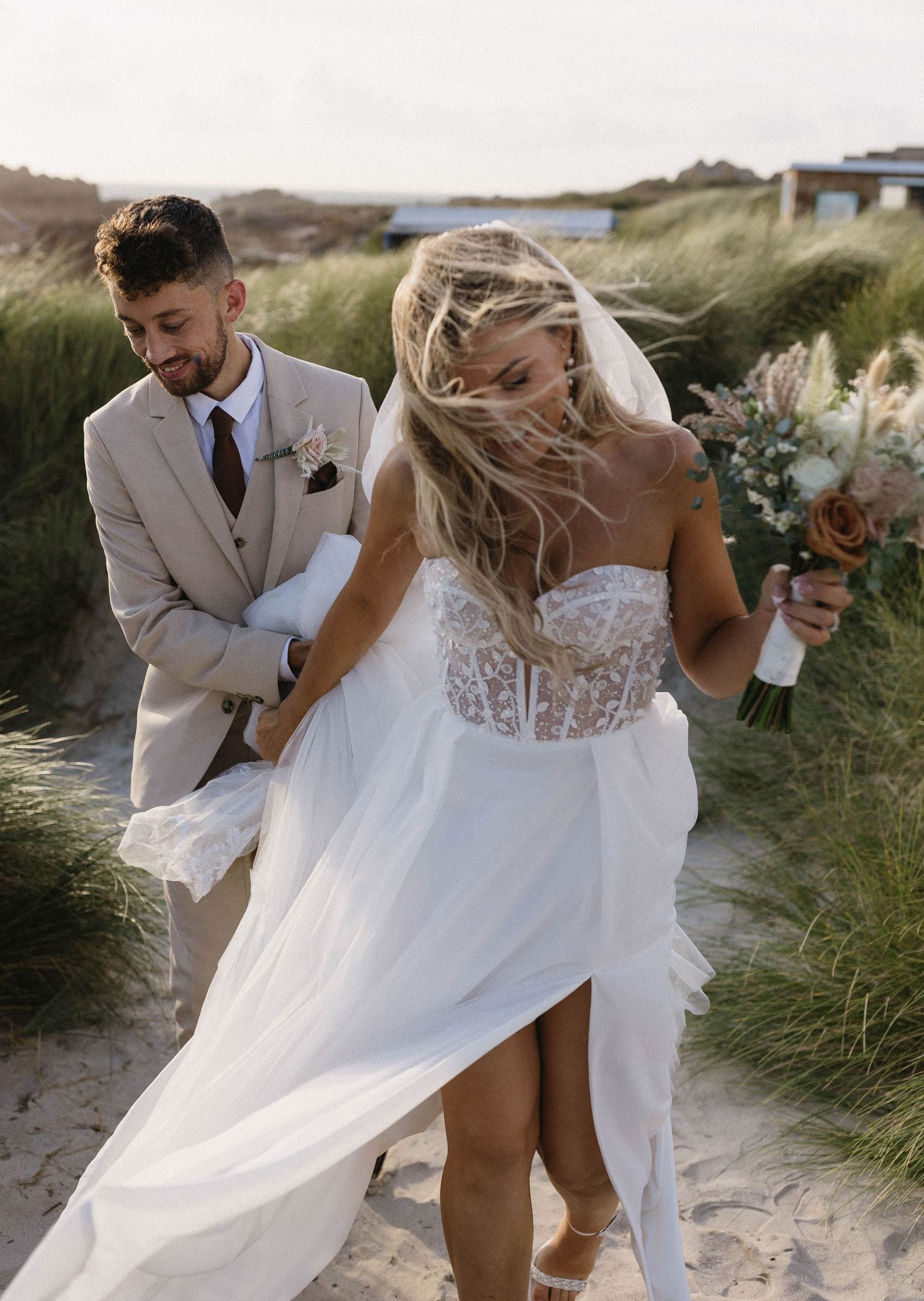 Couple in wedding attire playfully walking through beach grass on a windy day.