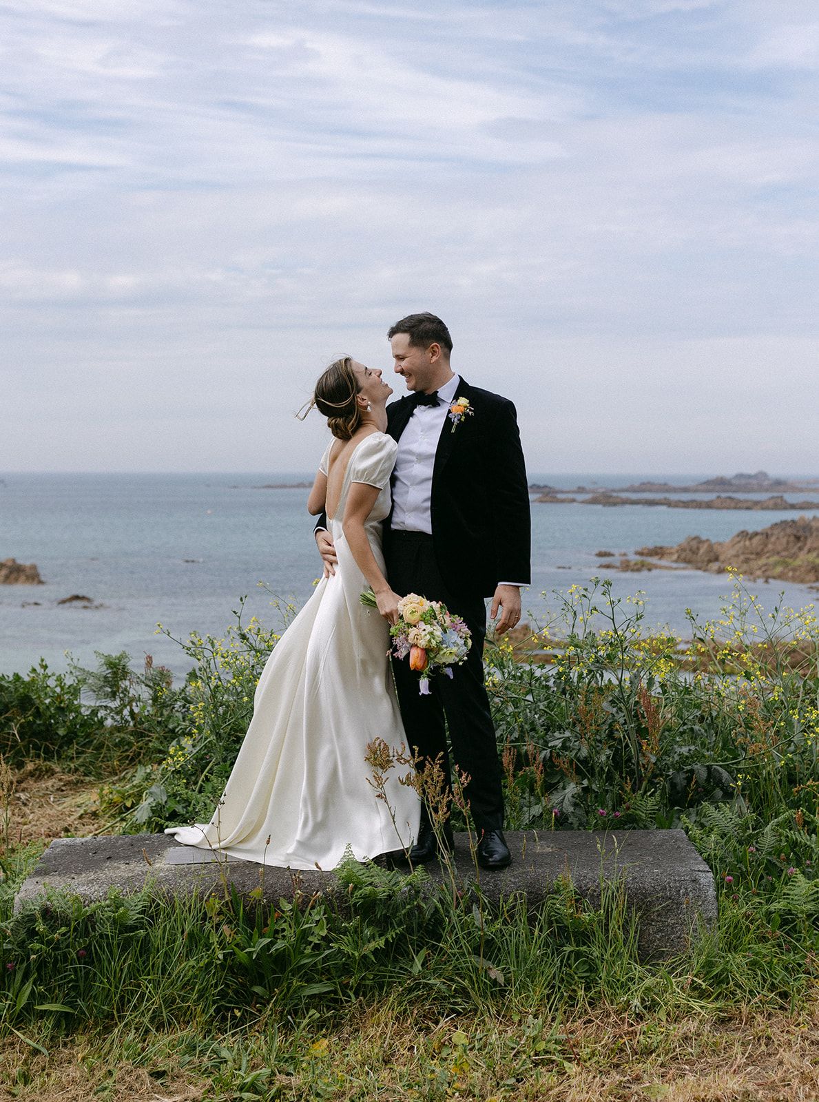 Bride and groom share a romantic kiss on a coastal cliff, with ocean views and wildflowers in the background.
