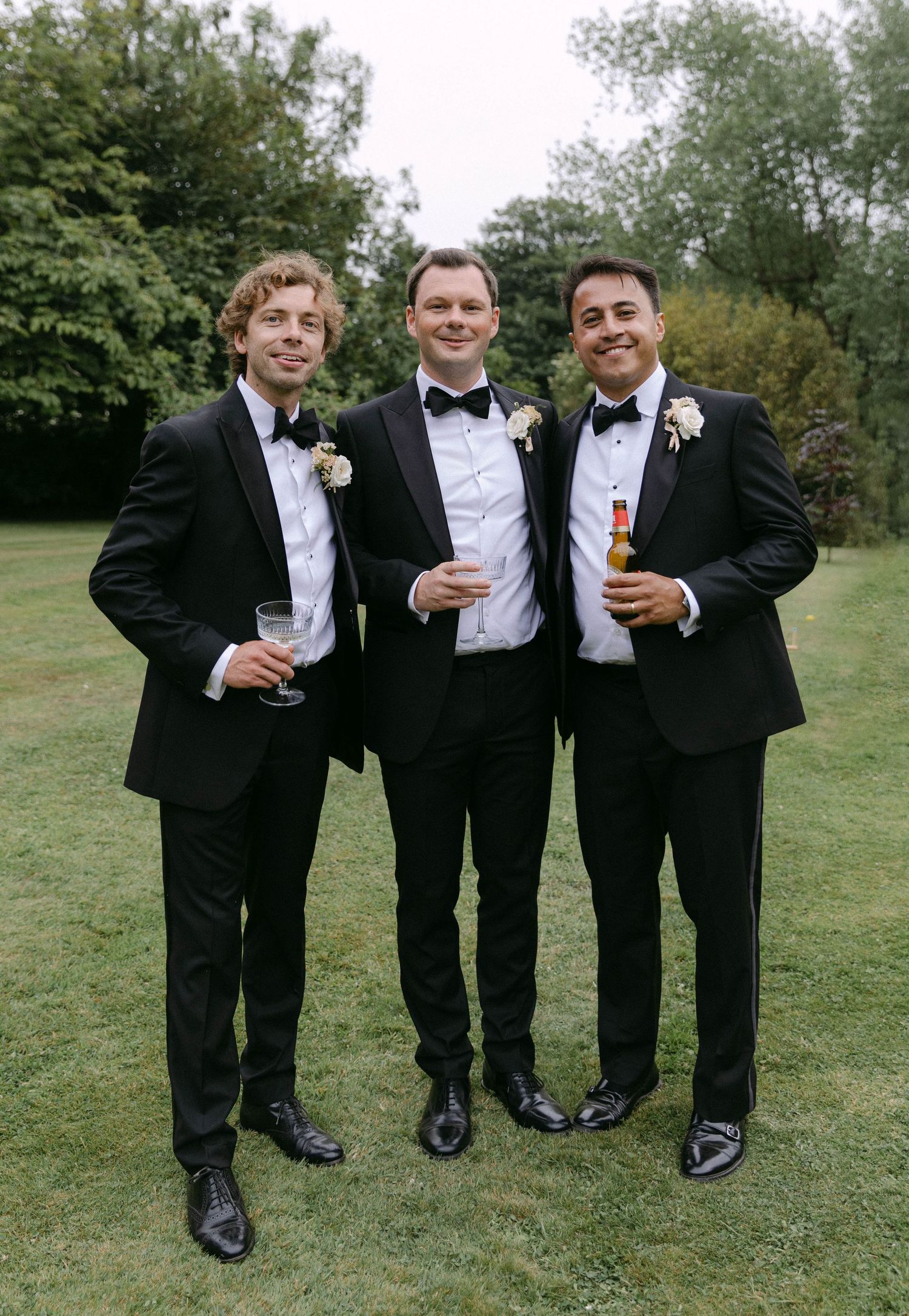 Three groomsmen in black tuxedos holding drinks and smiling on a lawn.