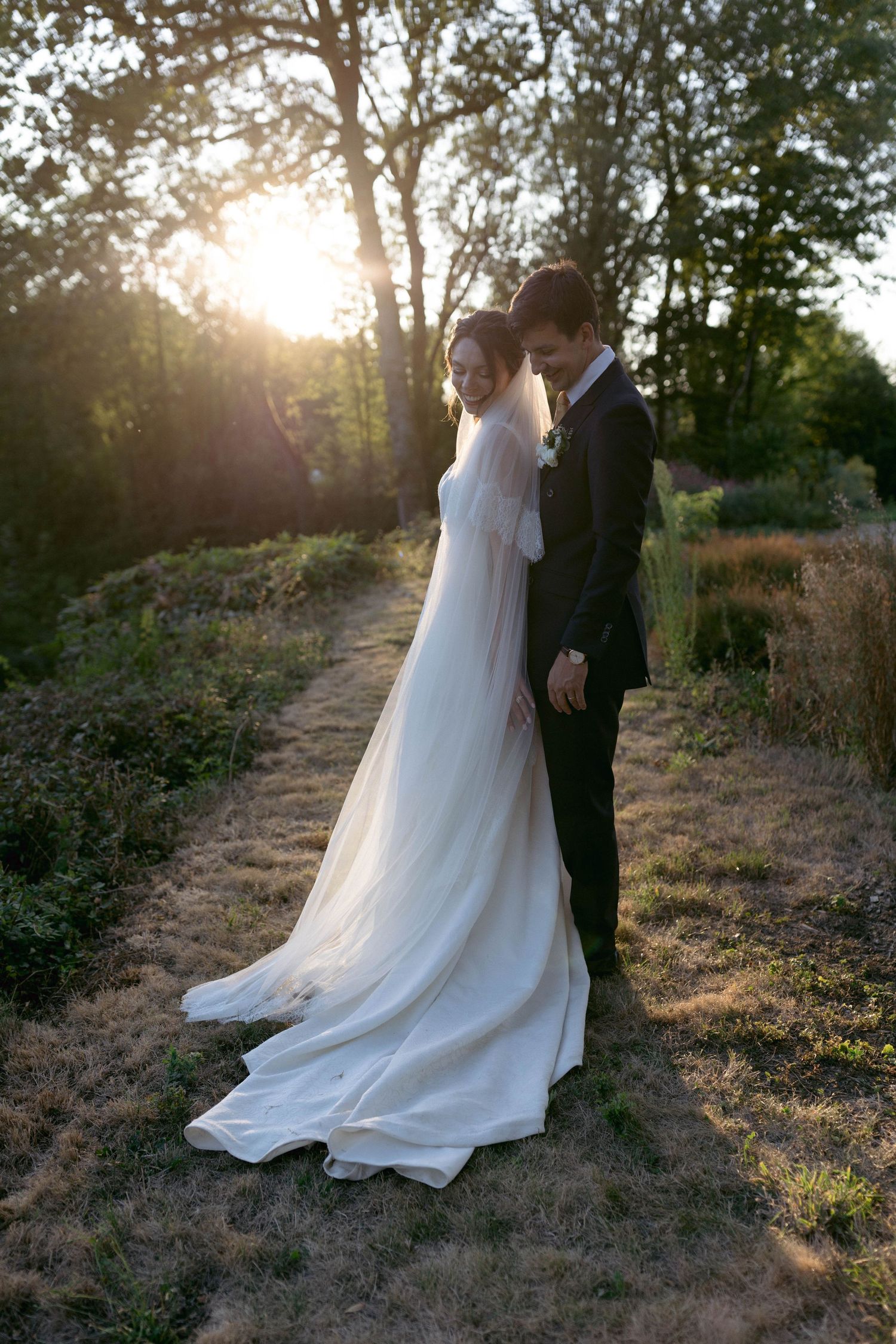 Bride in flowing white gown and veil stands with groom in dark suit at golden sunset in lush garden.