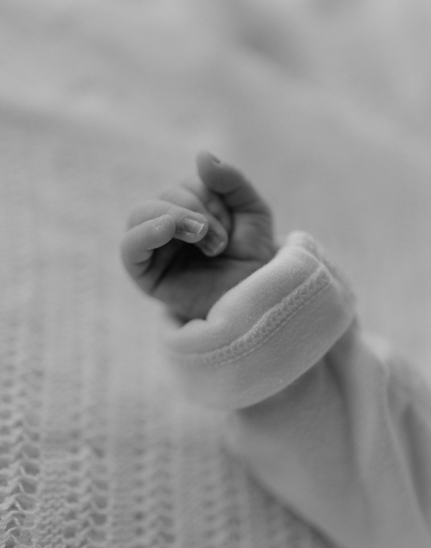 Close-up black and white photo of a newborn baby's tiny hand with delicate fingers resting on a soft white blanket.