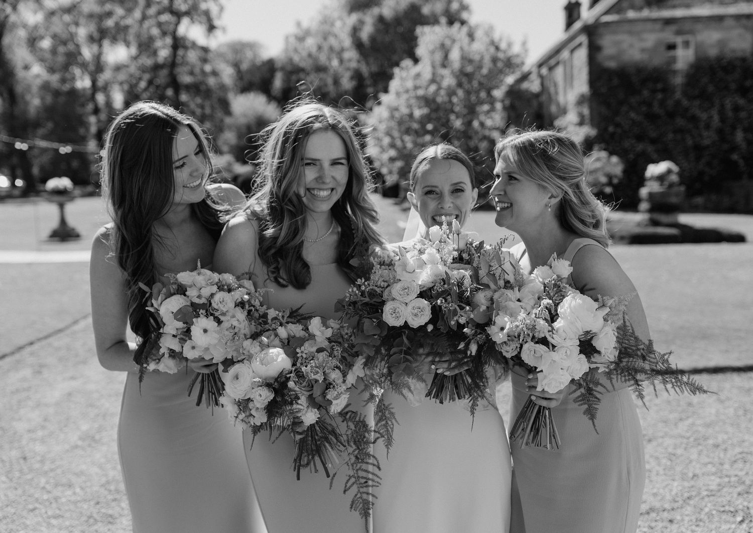 Bridesmaids in elegant dresses hold beautiful white and pink floral bouquets outdoors.