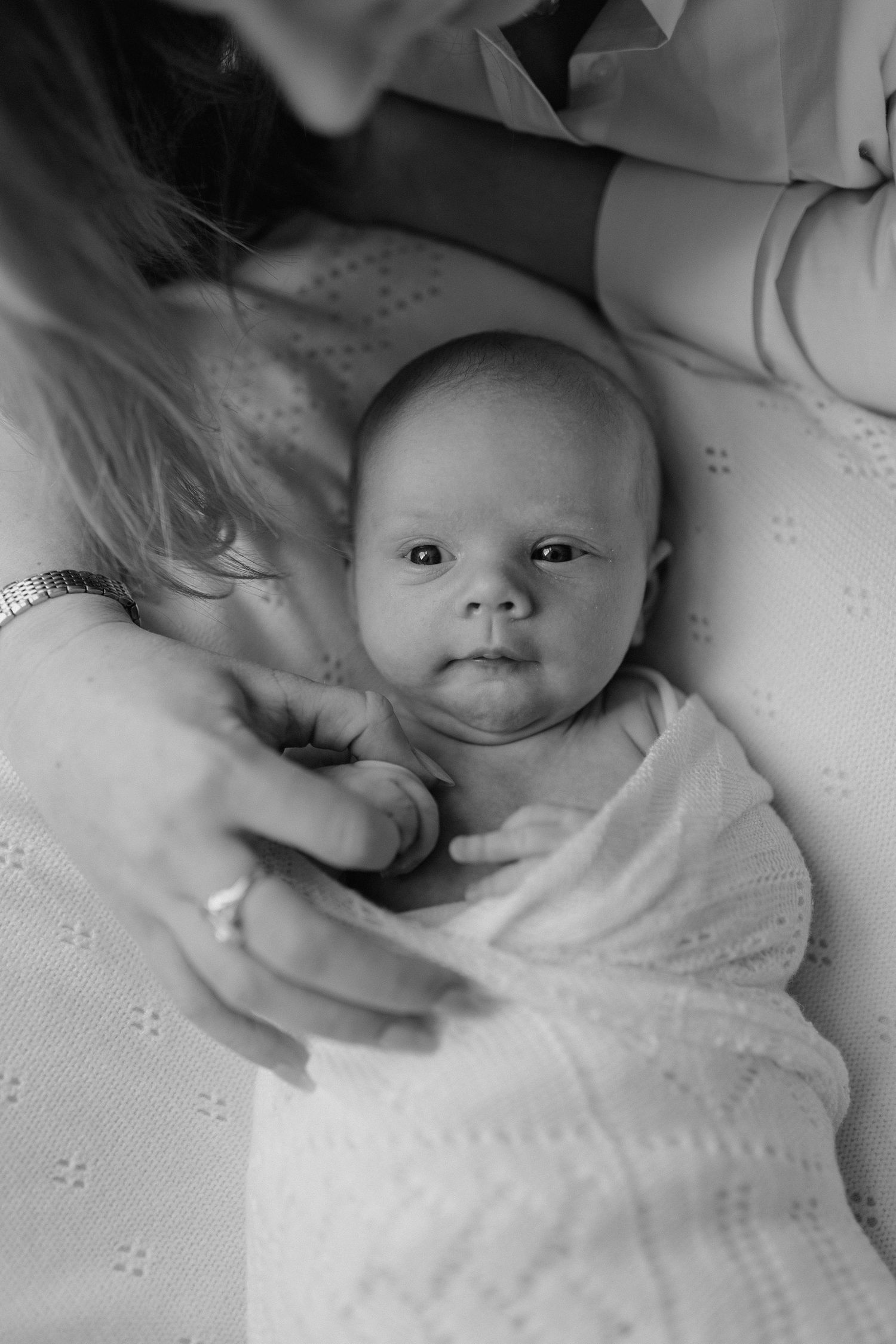 Black and white photo of a newborn baby swaddled in white, gazing up while a mother tenderly holds their tiny hand.