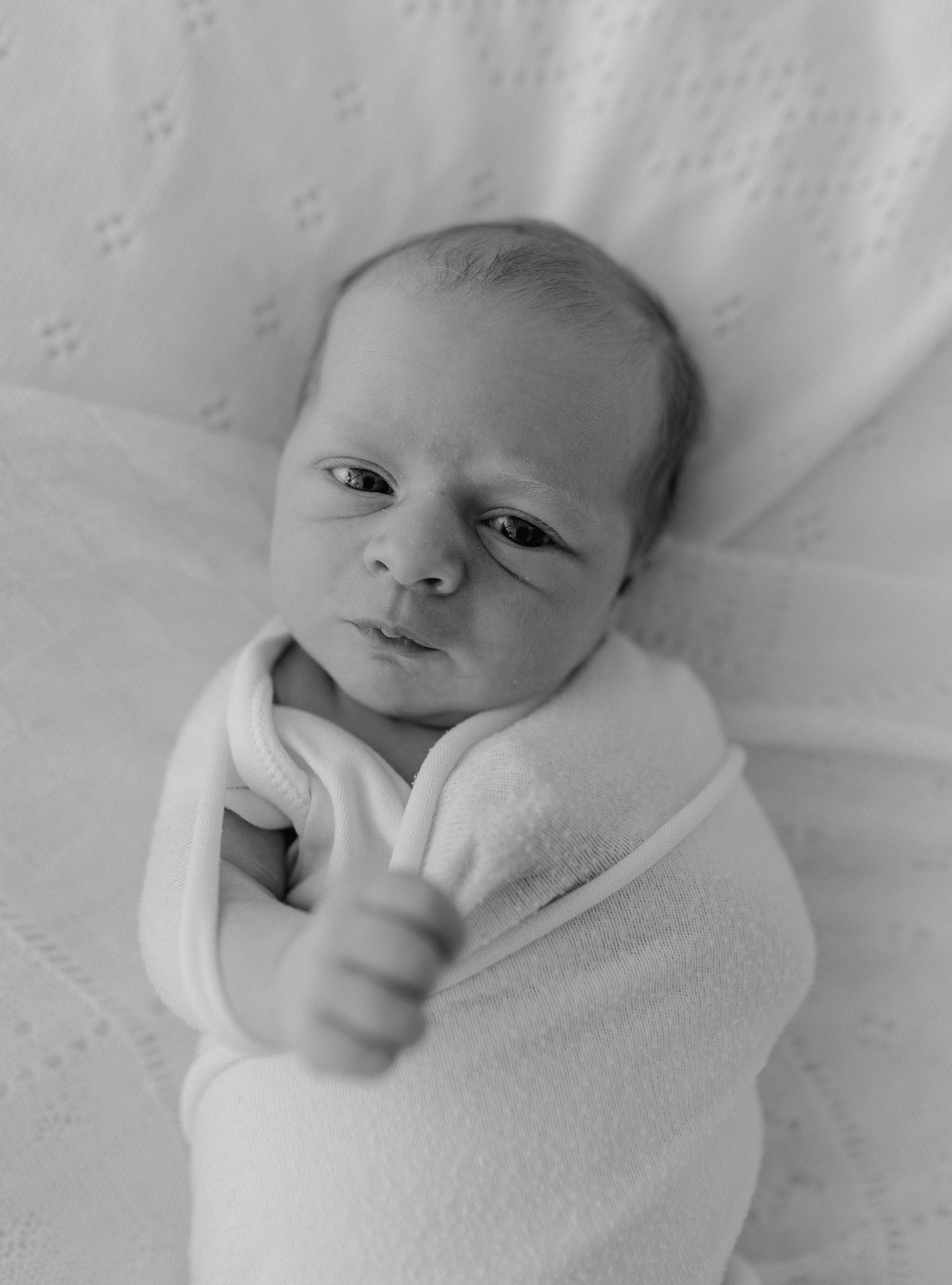 Black and white photo of a newborn baby swaddled in a white blanket, looking up with wide eyes.