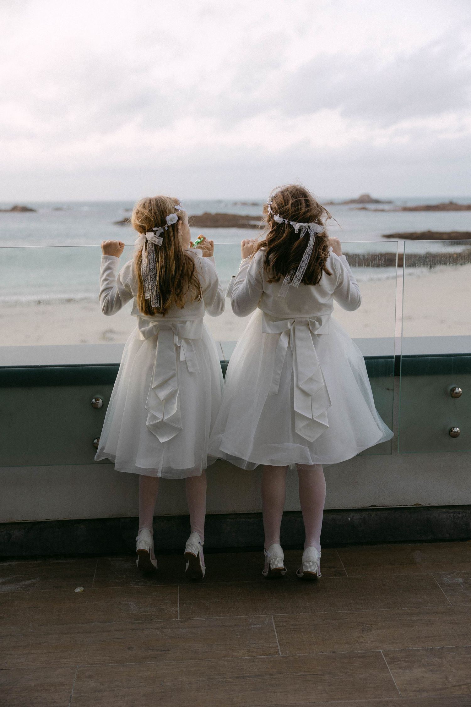 Two flower girls in matching white dresses overlooking ocean view from balcony.