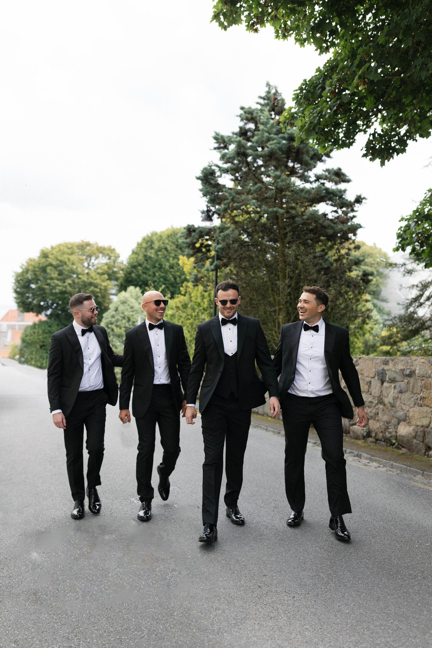 Groomsmen in classic black tuxedos walking together on a paved path surrounded by trees.