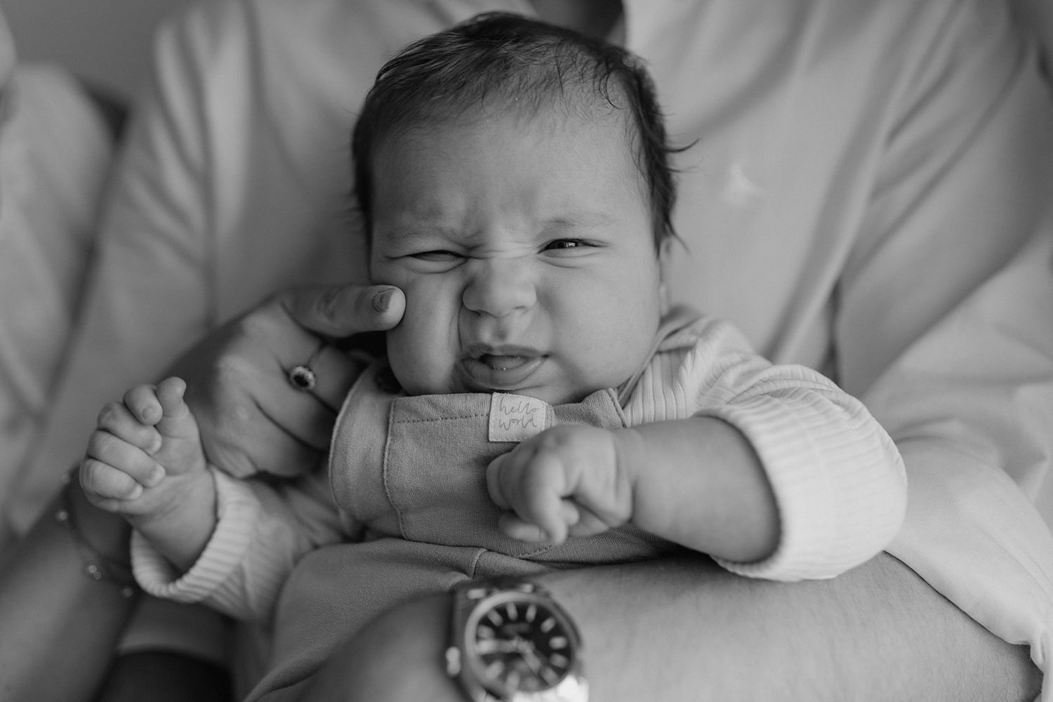 Black and white photo of a newborn baby making a funny face while being held by an adult wearing a watch.