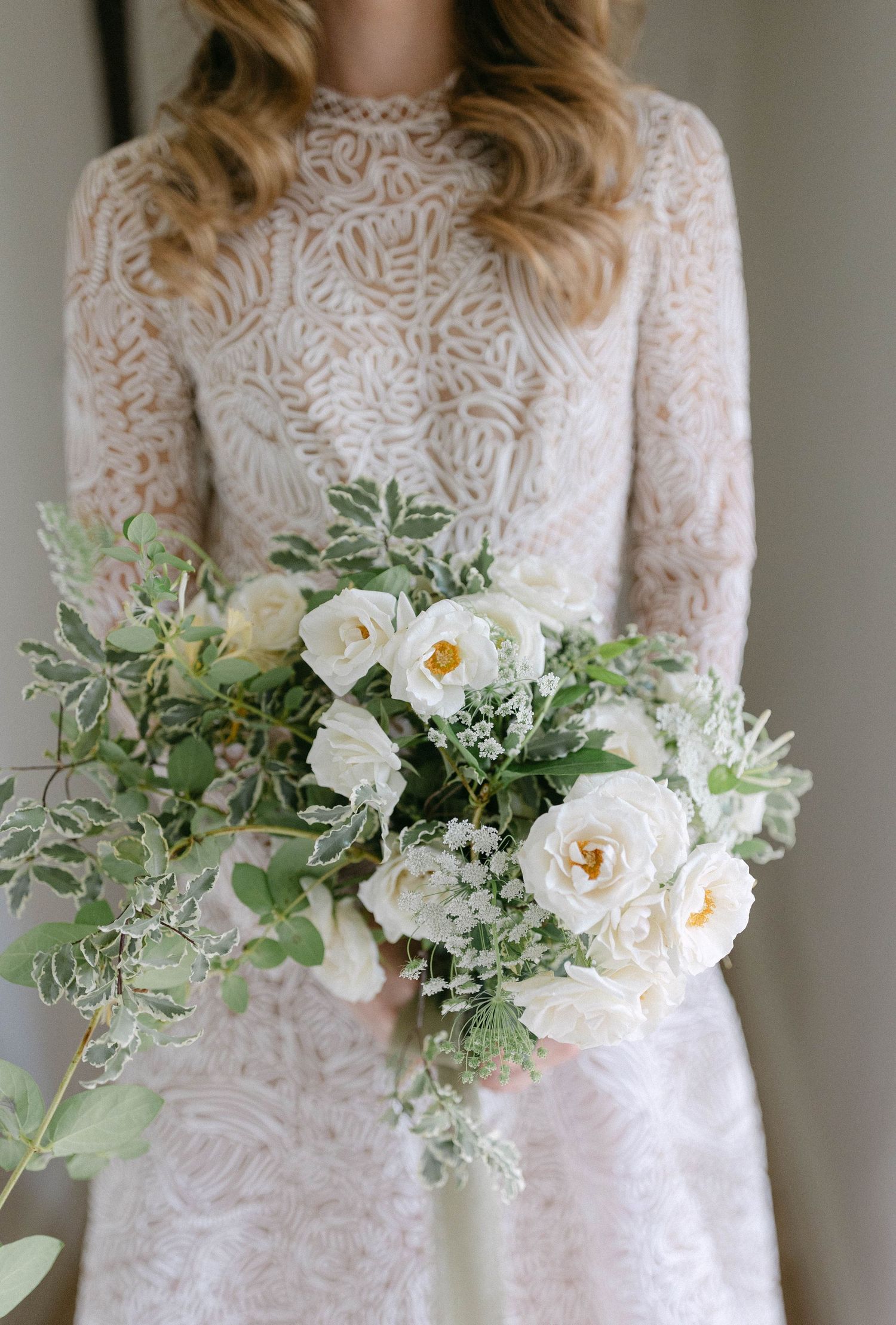 A delicate white bridal bouquet with roses and greenery held against a lace wedding dress.