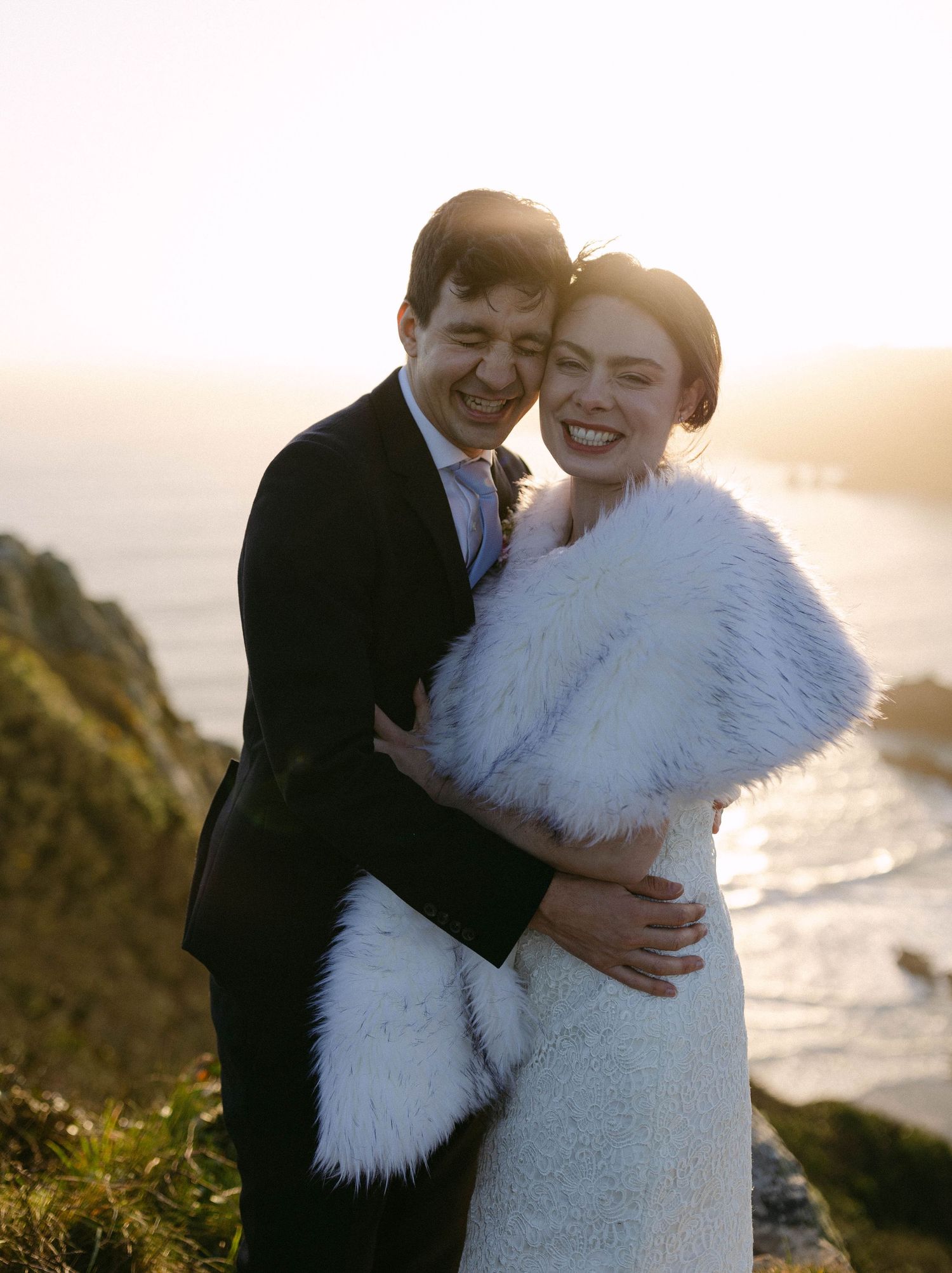 Romantic sunset wedding portrait on a coastal cliff with white fur wrap and ocean backdrop.