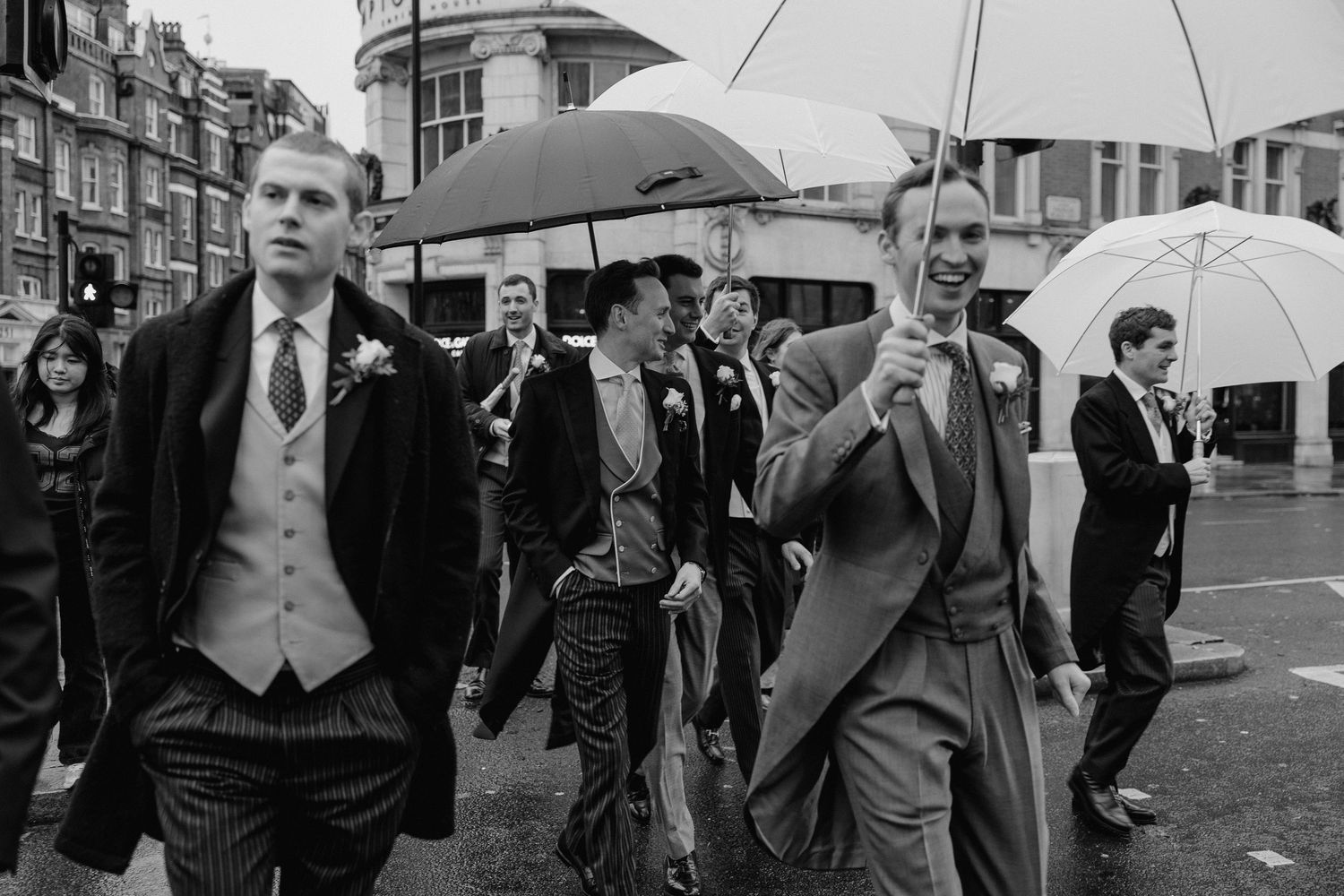 Wedding guests gather outside on a rainy day holding umbrellas in formal attire.