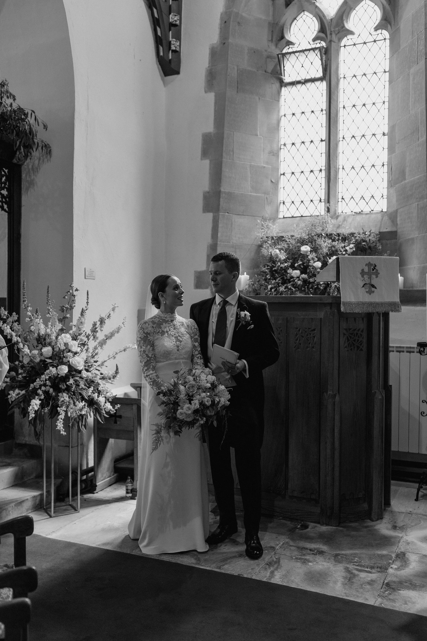 A wedding ceremony in a church with gothic windows and floral arrangements.