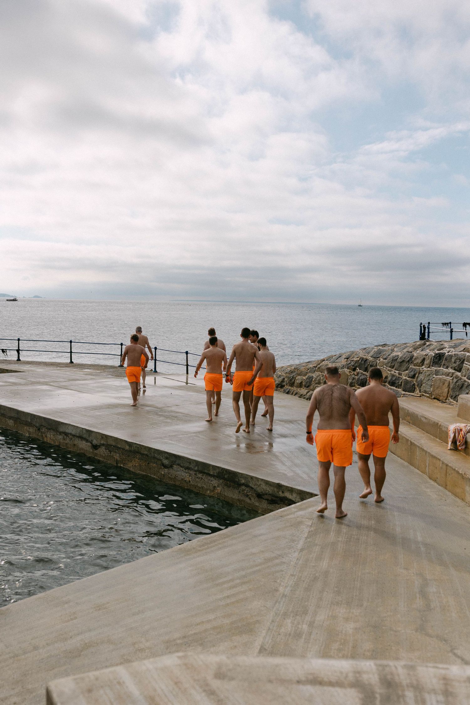 People in orange swimwear walk along a concrete pier by the ocean. Bathing Pools Guernsey.