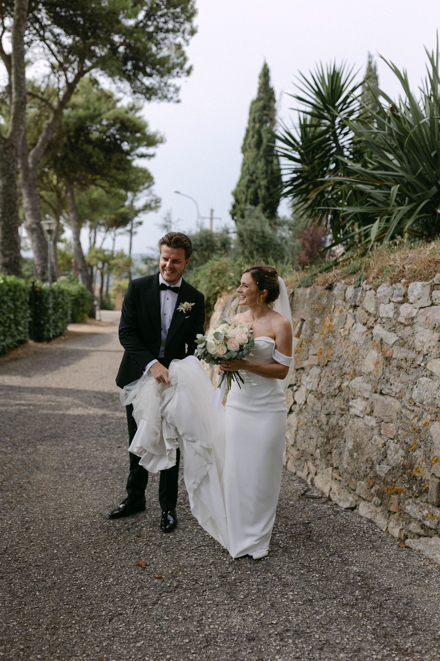 Wedding couple seated on stone wall surrounded by Mediterranean cypress trees.