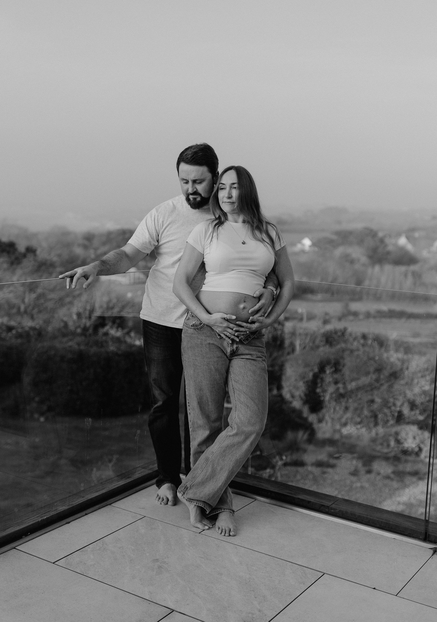 Black and white photo of a couple on a balcony, woman showing pregnant belly, scenic landscape background.