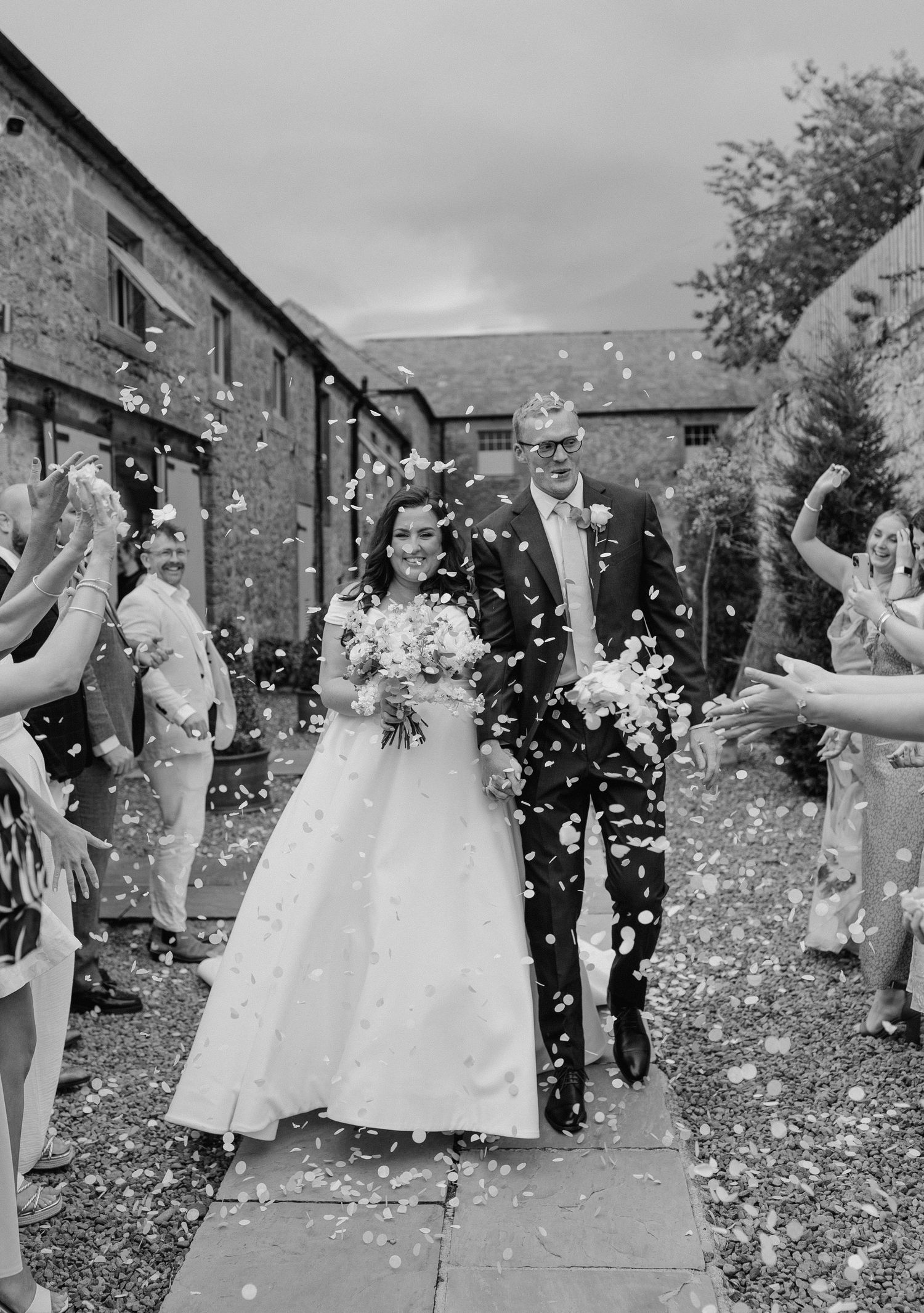 Bride and groom walk through confetti shower, guests lining path, barn wedding venue, black and white photo.