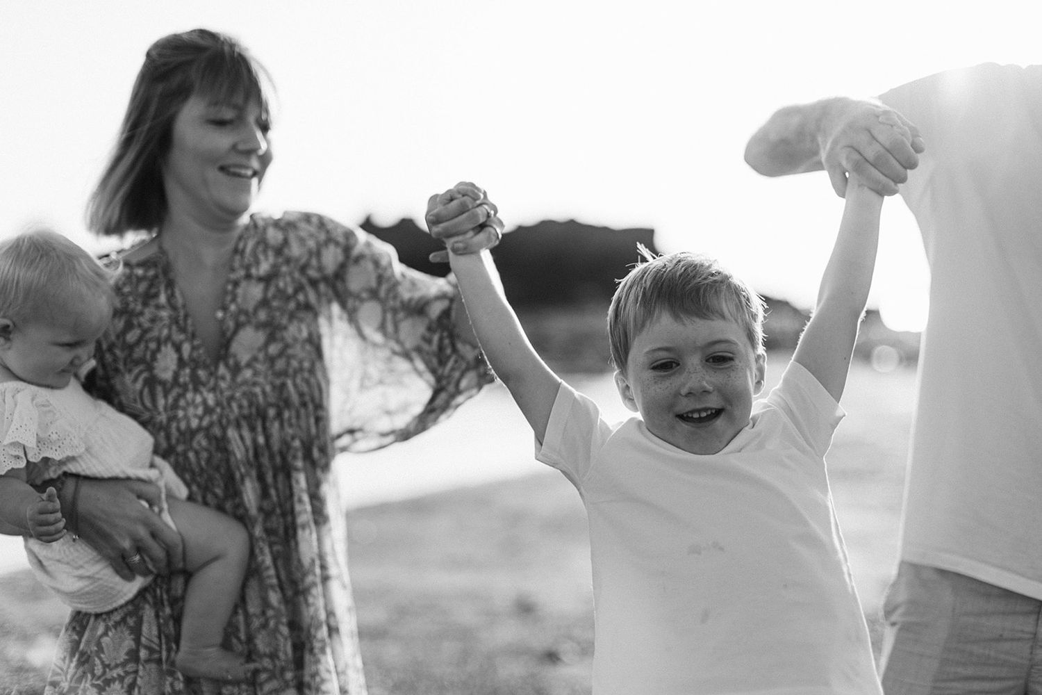 Black and white photo of a joyful family at the beach, mother holding baby while child raises arms with delight.