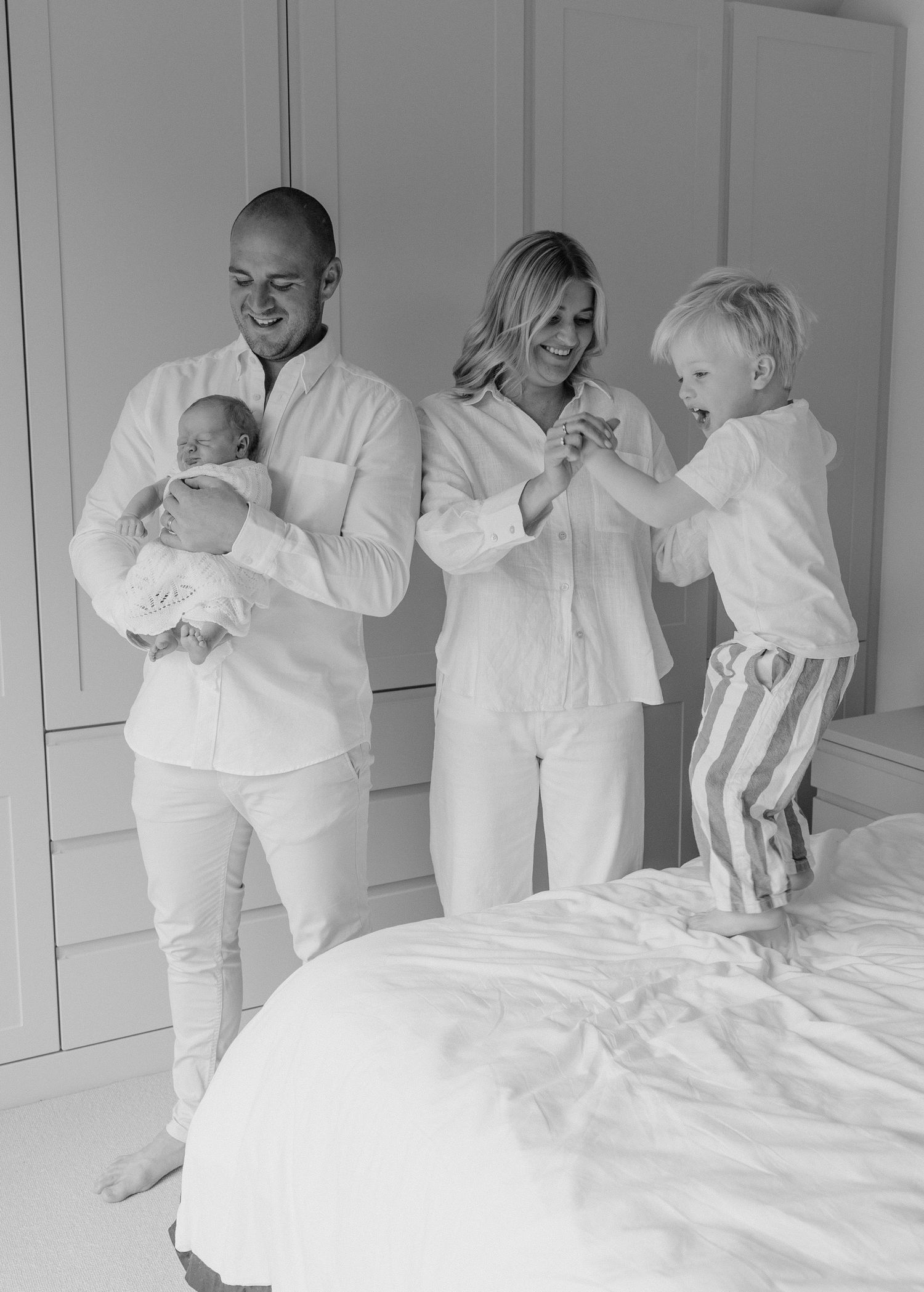 Black and white family photo of parents with newborn baby and toddler standing on bed in white bedroom.