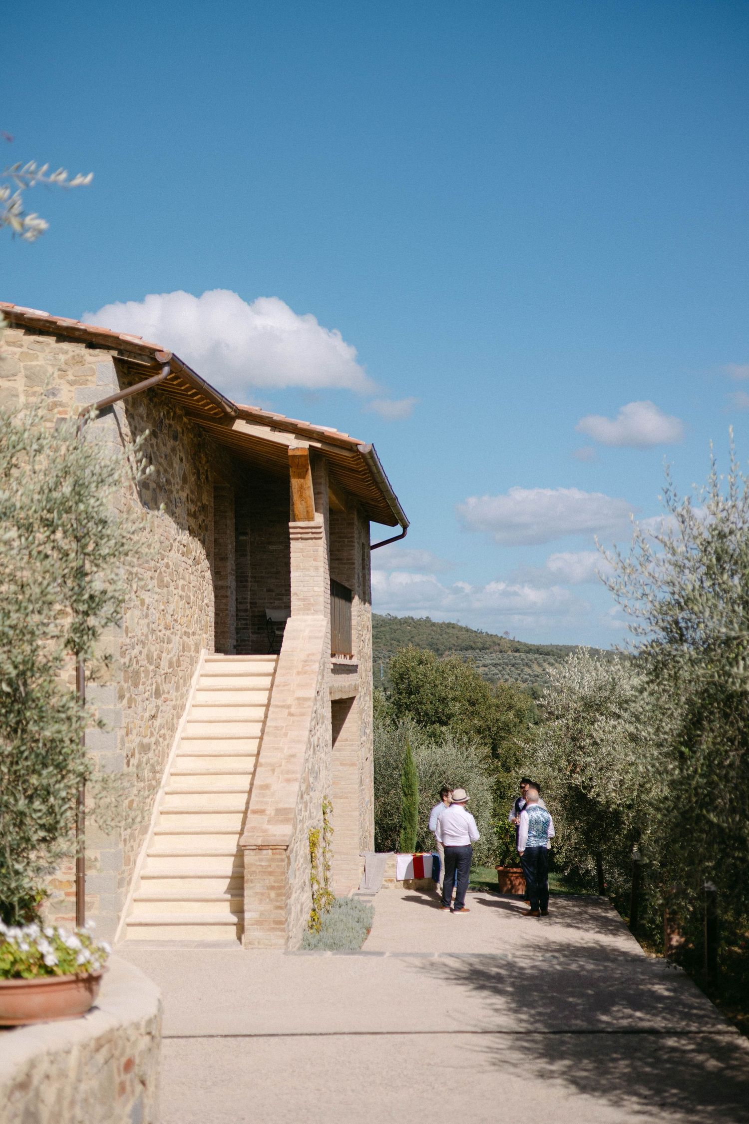 Two people walk along a stone path leading to a rustic Tuscan villa on a sunny day.