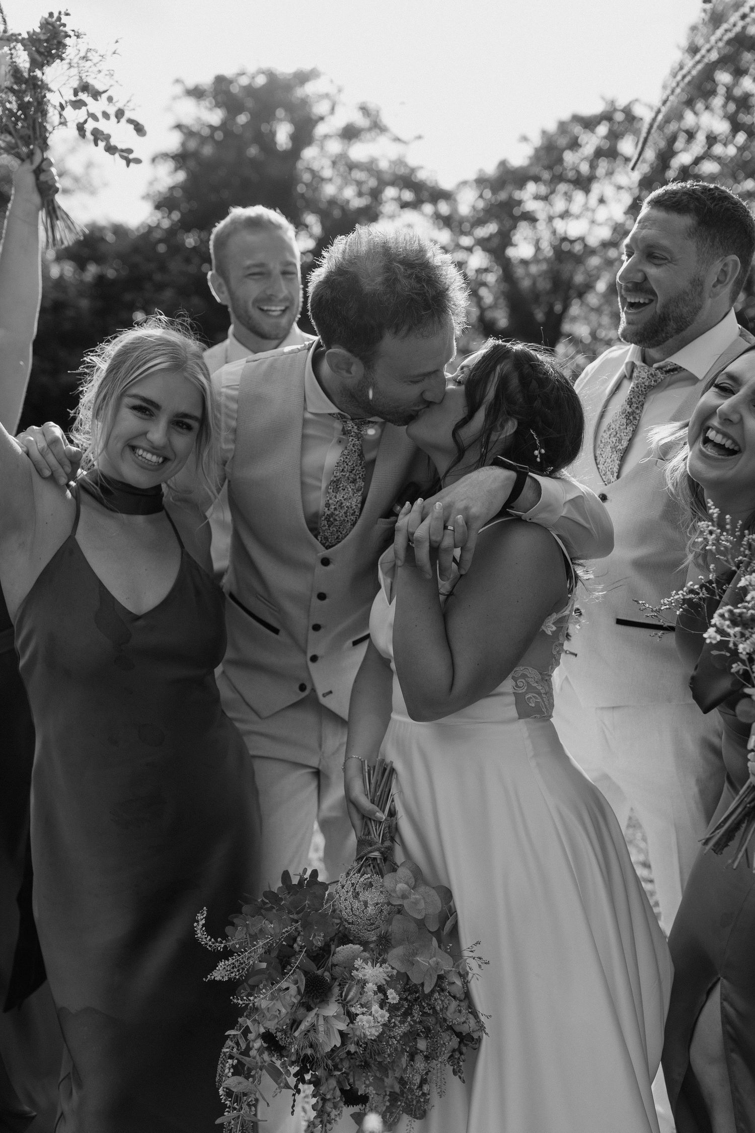 A wedding party laughs together outdoors in a candid black and white photo.