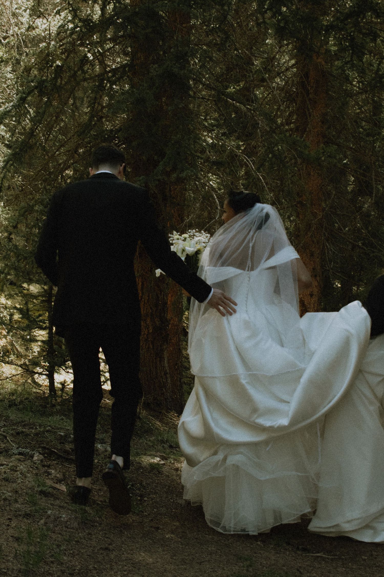 Groom in black suit guides bride in flowing white gown and veil through a lush forest setting.
