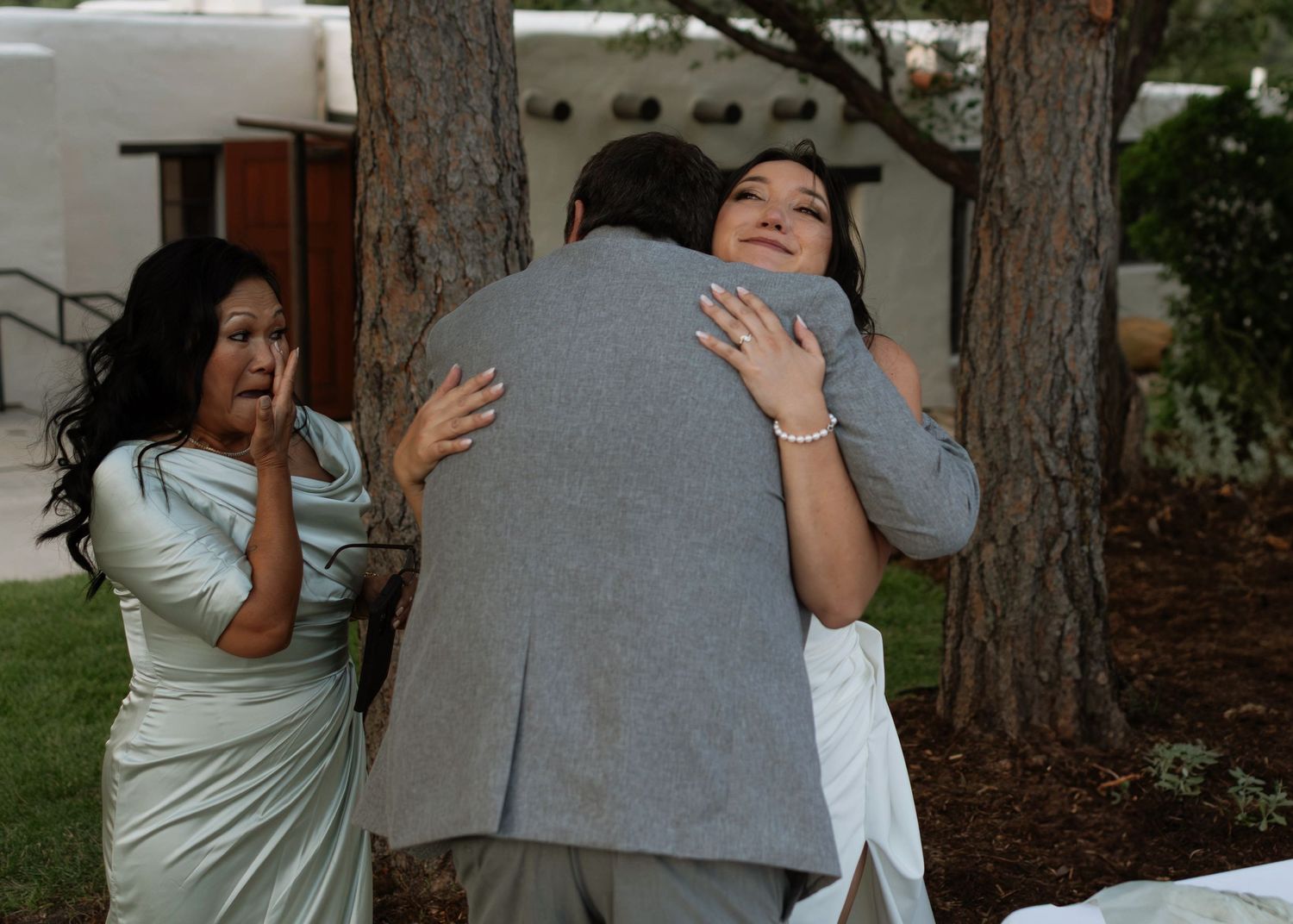 Man in gray suit hugging woman in white dress at outdoor wedding while another woman in green watches nearby.
