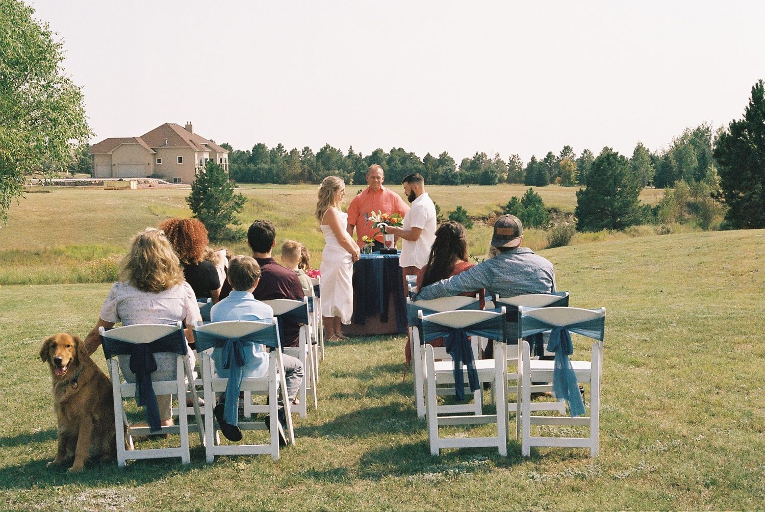 Outdoor wedding ceremony on green field with white chairs, guests seated, officiant at altar, dog beside attendees.