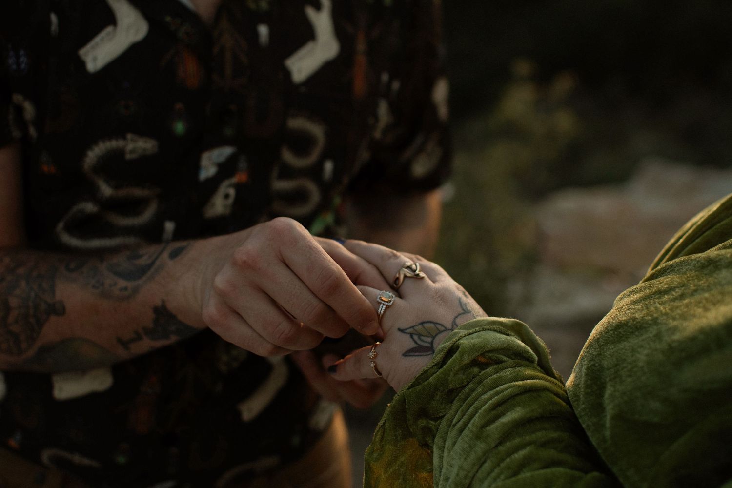 Tattooed hands holding a small object outdoors, wearing a patterned dark shirt with snake designs.