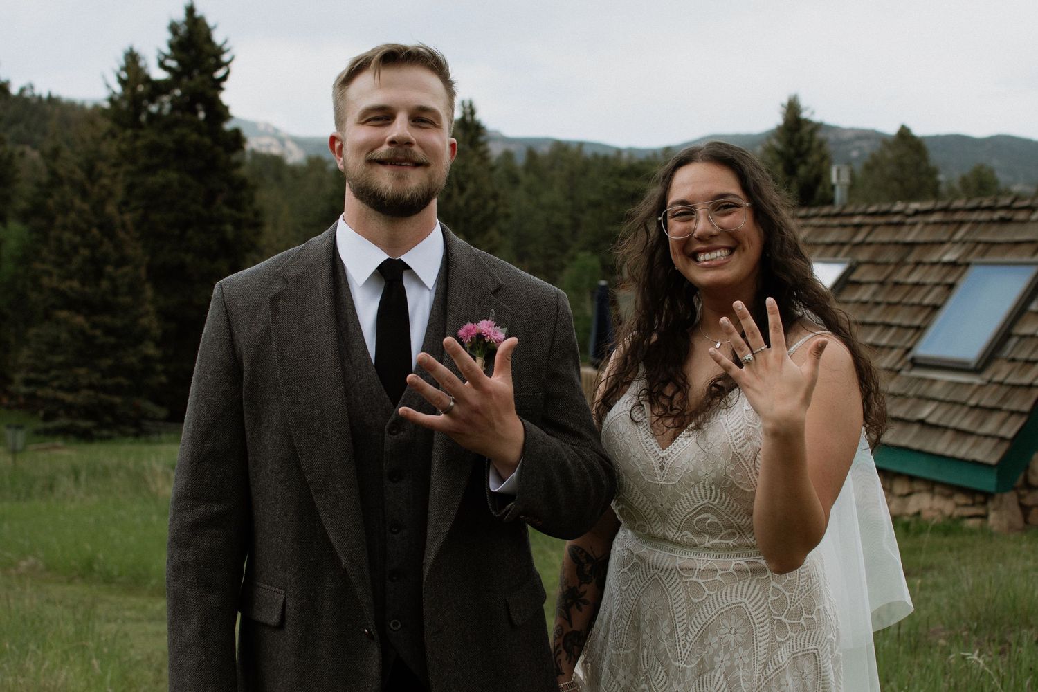 Newlywed couple smiling and showing wedding rings outdoors with mountain forest backdrop.