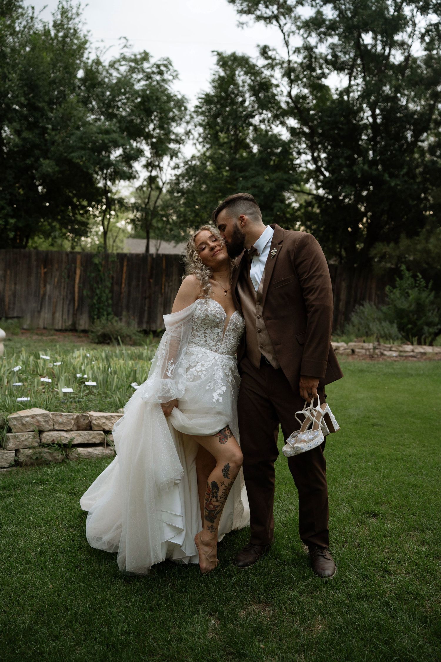 Bride and groom share a romantic kiss in a lush garden, bride in lace gown with tattoos, groom in brown suit.