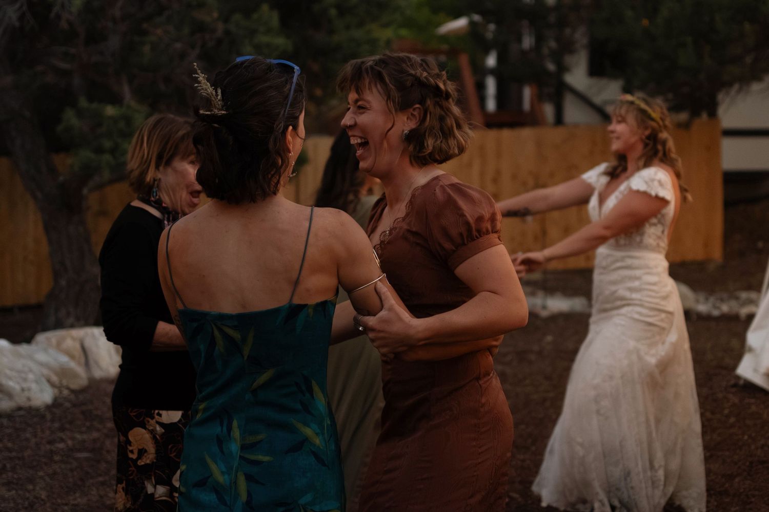 Women dancing outdoors at a wedding celebration, with a bride in white dress visible in the background.
