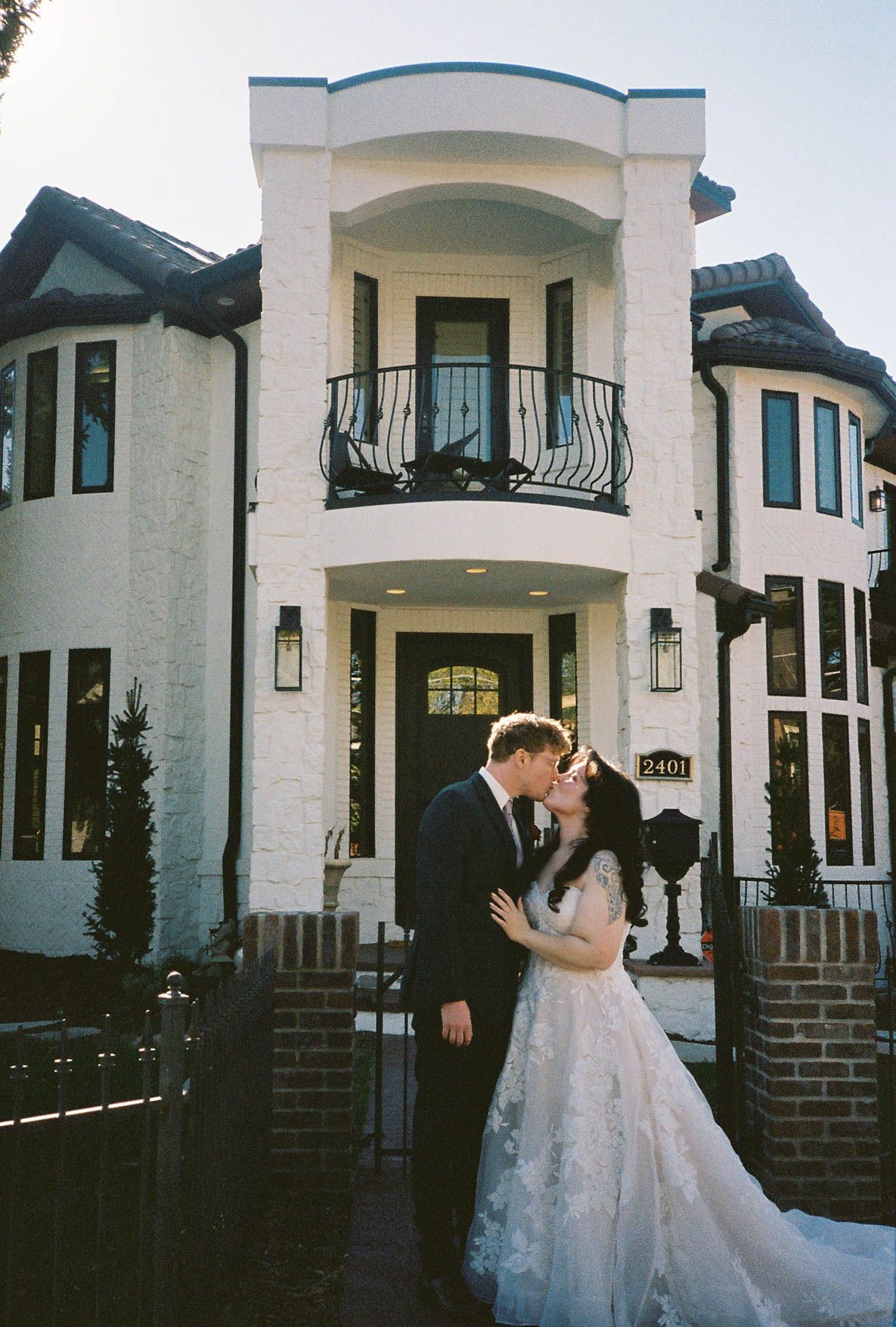 Bride and groom share a romantic kiss in front of an elegant white brick mansion with black iron balcony.