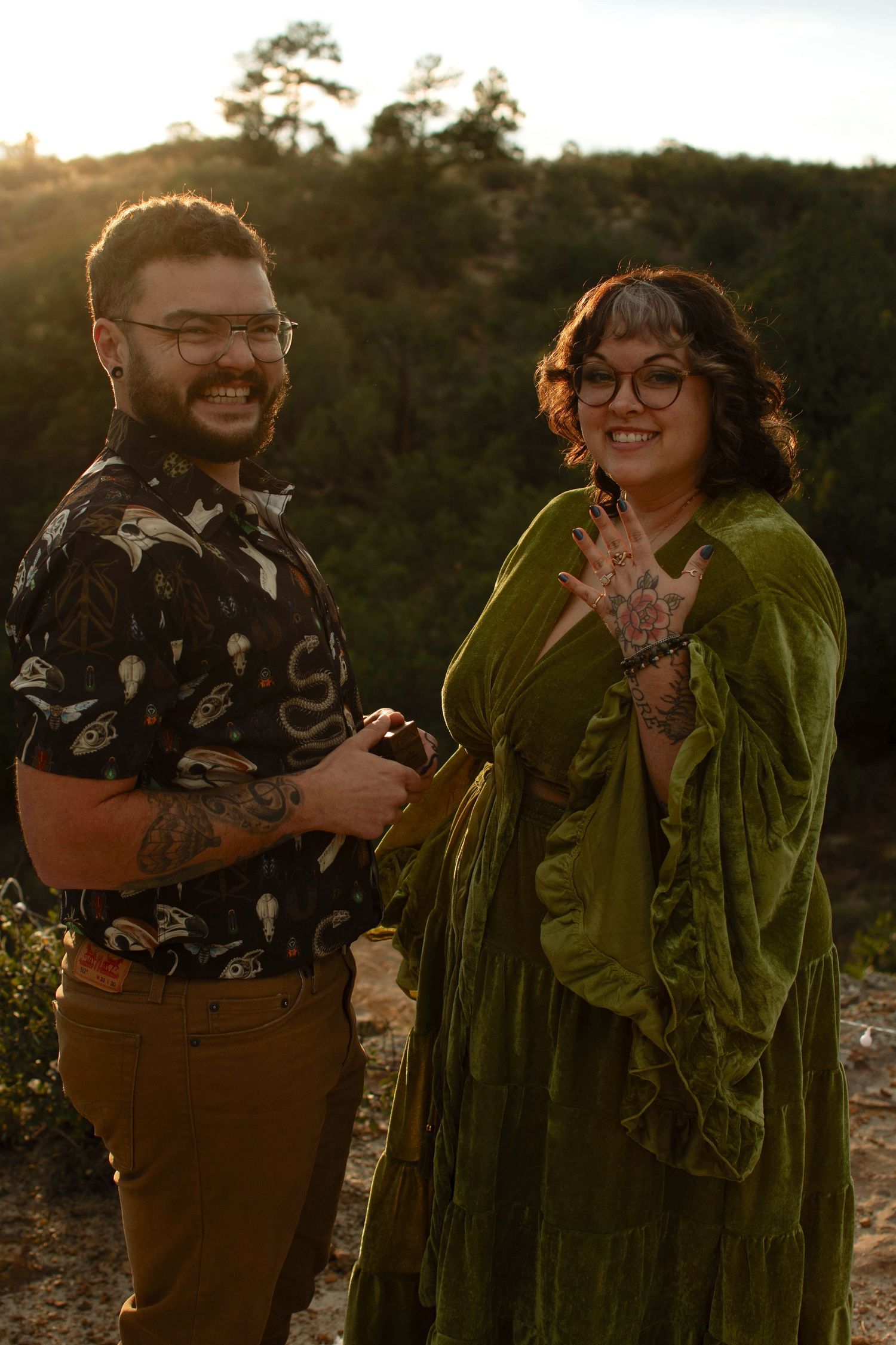 A smiling couple with tattoos and glasses poses outdoors at golden hour, the woman wearing a green velvet dress showing her ring.