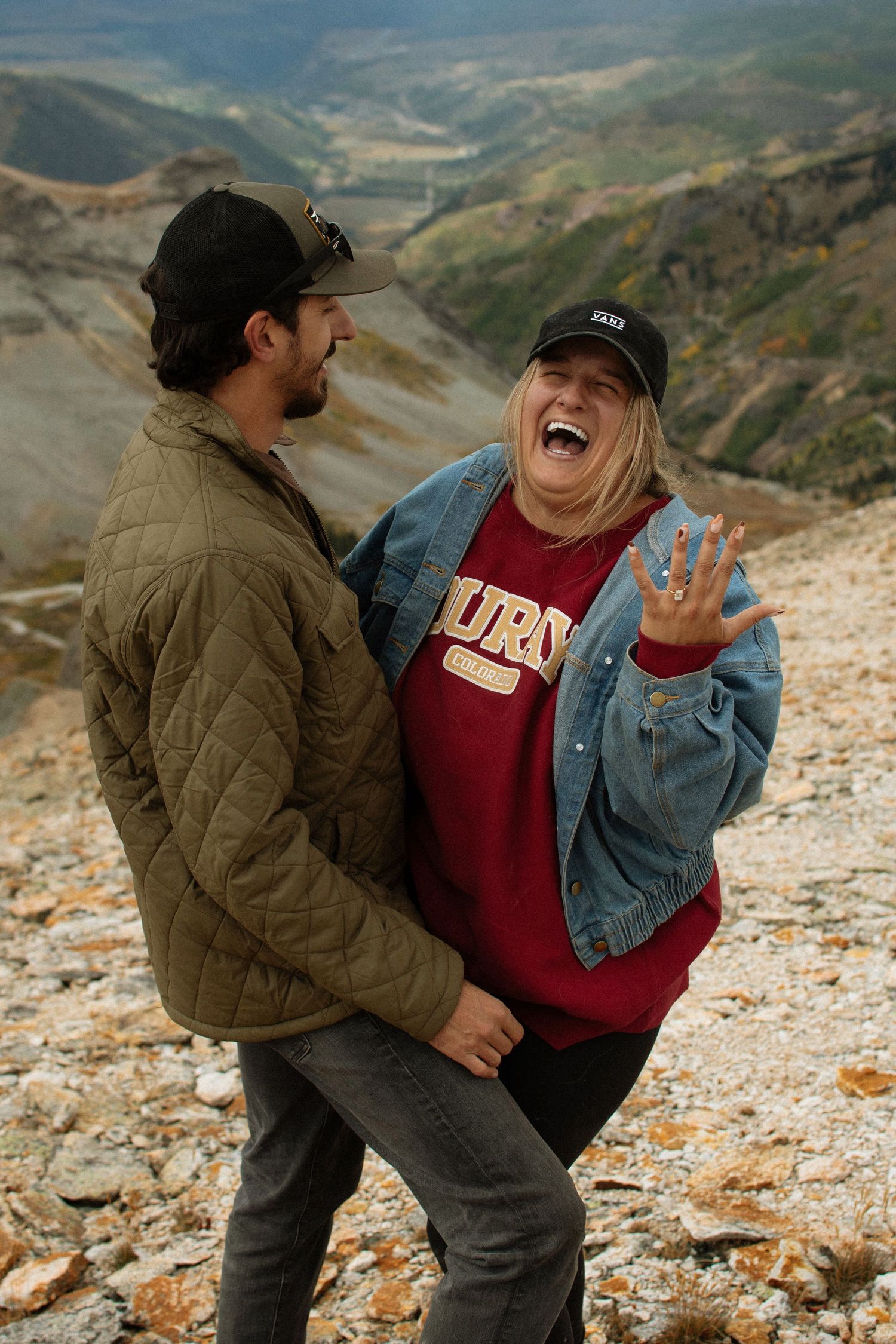 Couple laughing on a rocky mountain overlook, woman showing off engagement ring while wearing a burgundy sweatshirt.